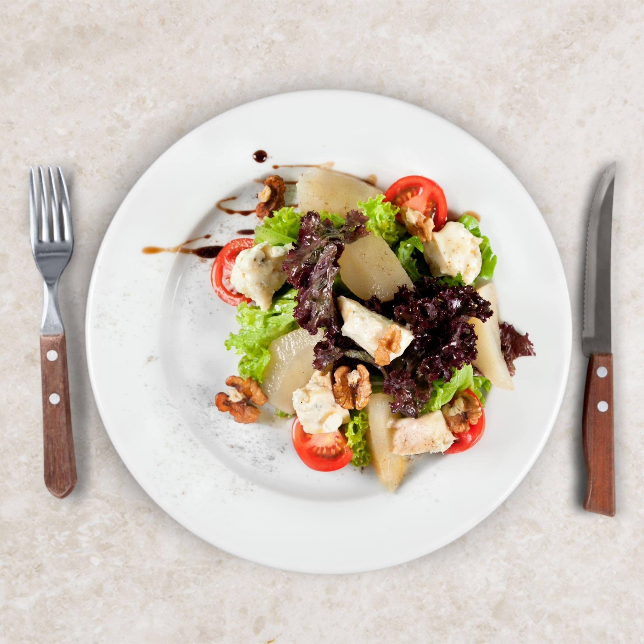 Ein Salat mit Birnen, Tomaten, Salat, Walnüssen und Käse auf einem weißen Teller mit Gabel und Messer.