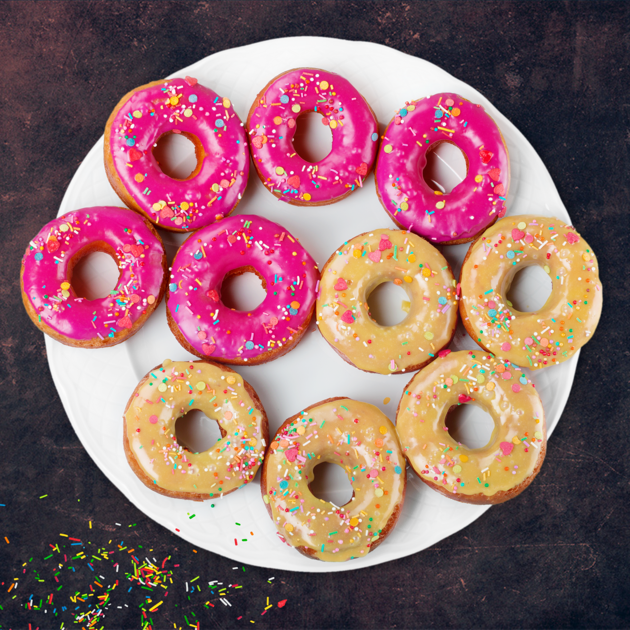 Draufsicht auf Donuts auf einem weißen Teller. Einige sind rosa und einige gelb, mit Streuseln verziert.