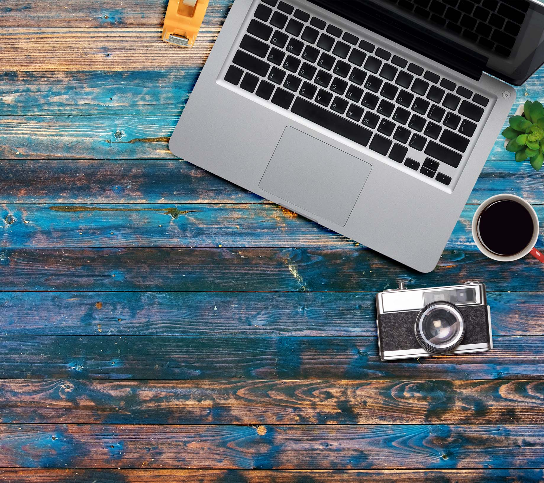 Ein Arbeitsplatz mit Laptop, Kamera, Kaffeetasse und Sukkulente auf blauer Holzoberfläche.