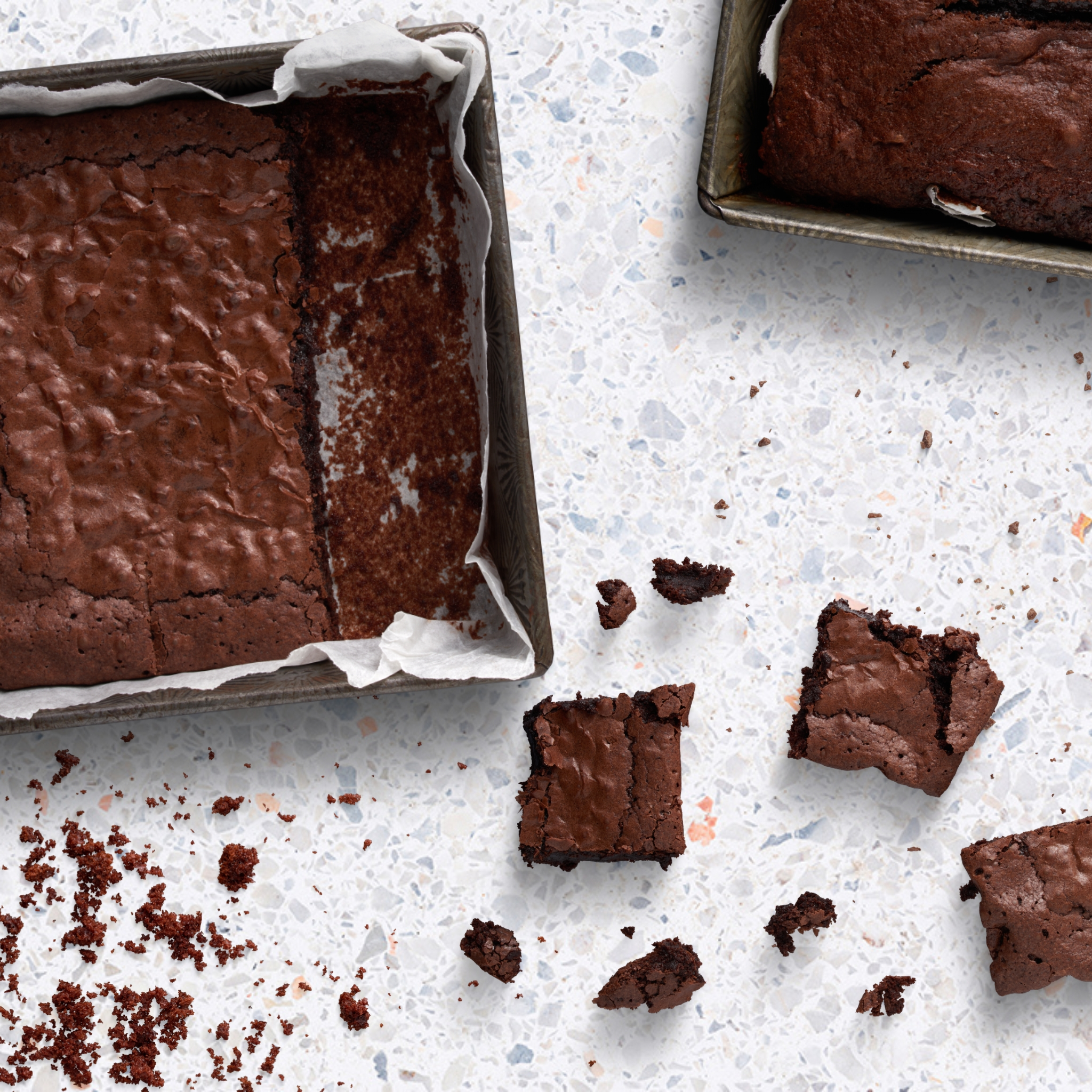 Brownies in einer Pfanne, mit einigen Stücken und Krümeln auf einer weißen, gesprenkelten Oberfläche.