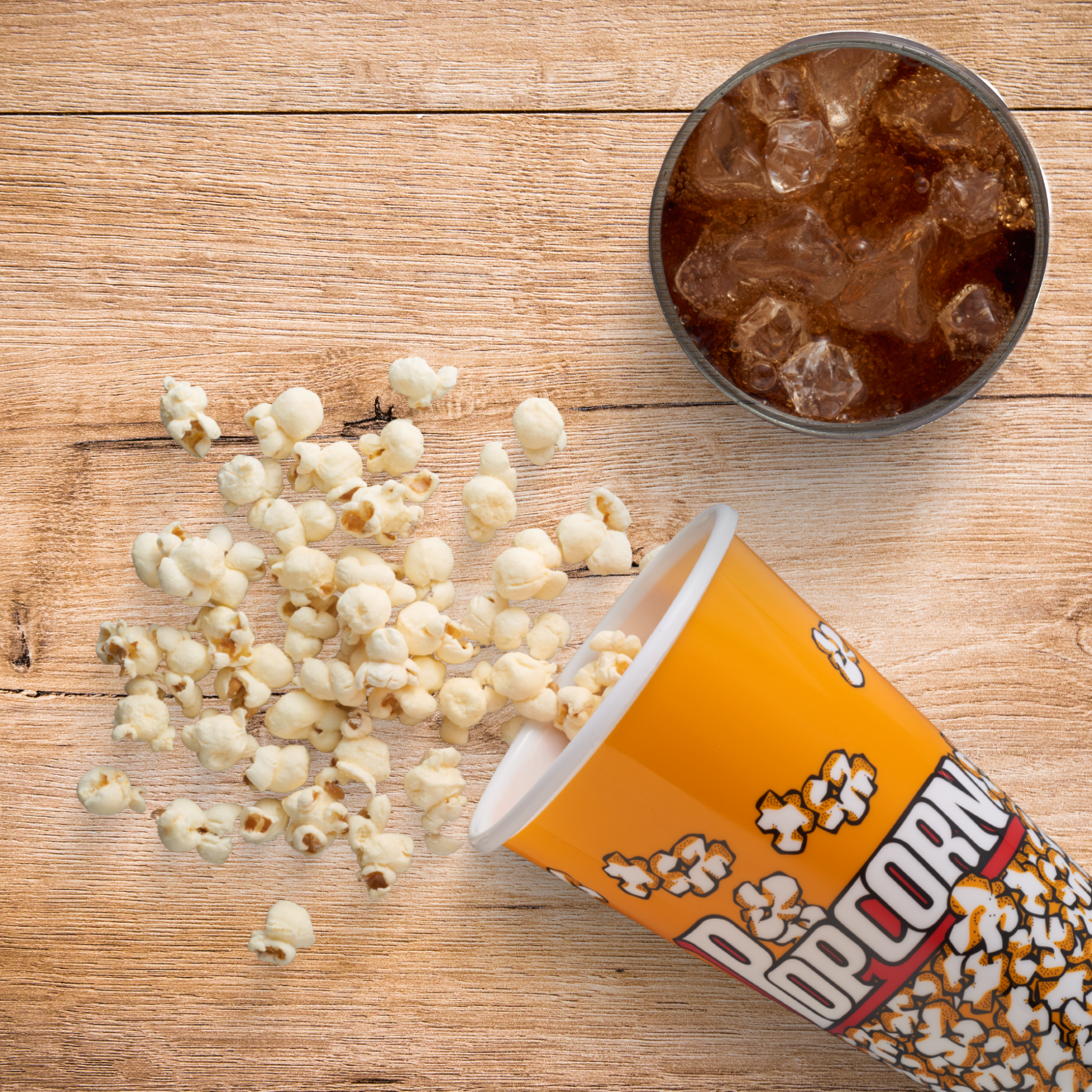 Popcorn quillt aus einem gelben Becher. Ein Glas Cola mit Eis steht auf einem Holztisch.