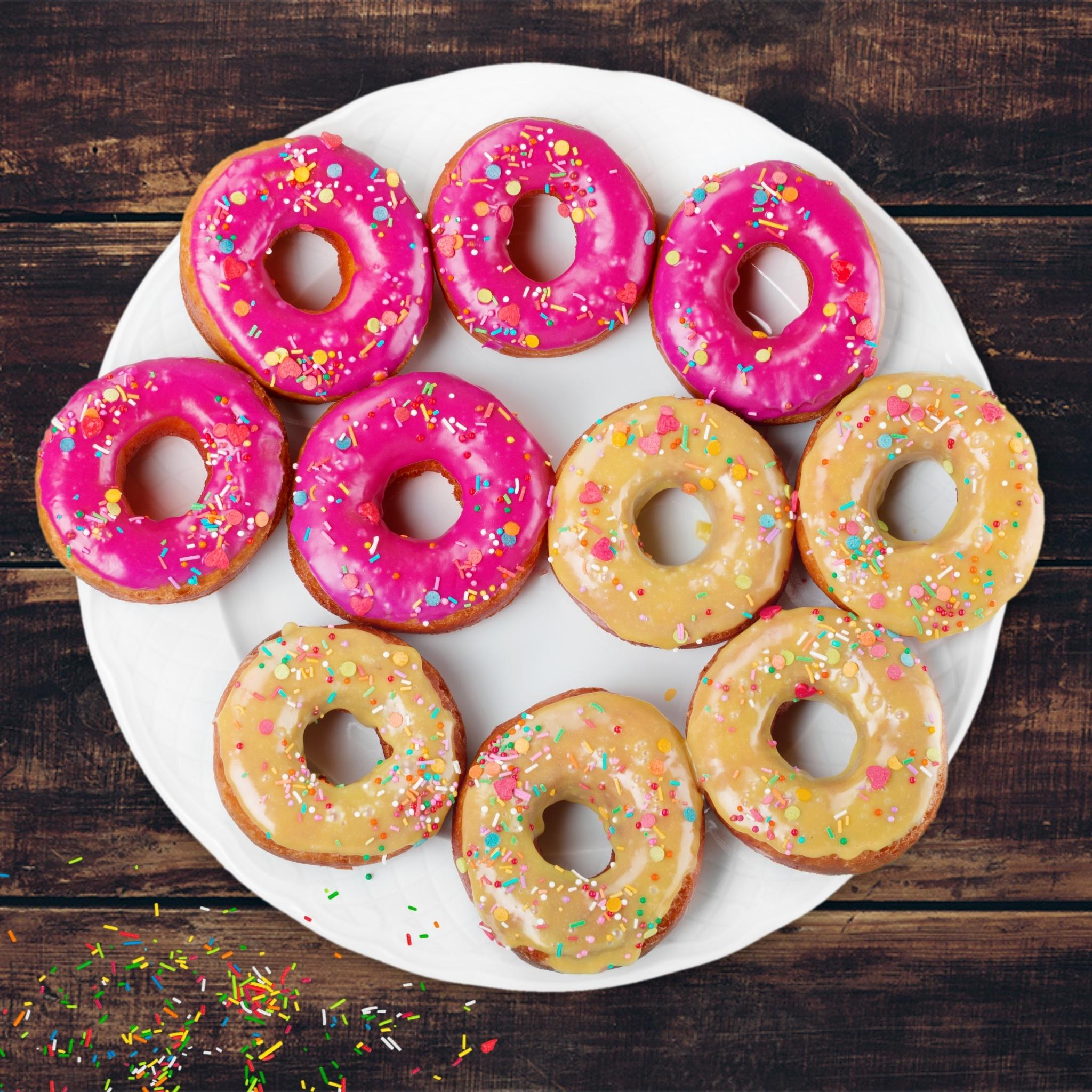 Ein weißer Teller mit Donuts, rosa und gelber Glasur, Streuseln oben drauf. Dunkler Holzhintergrund.