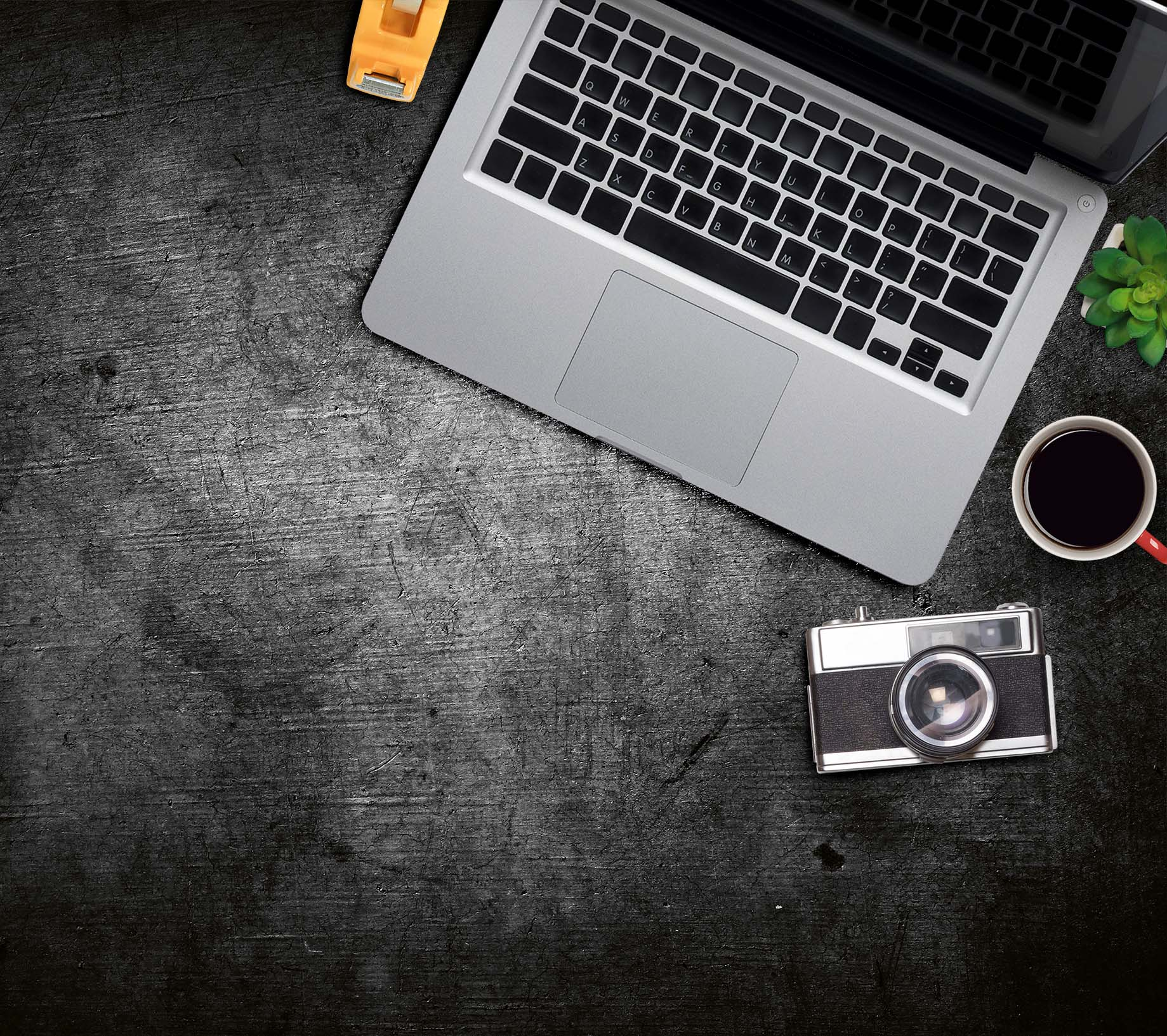 Schreibtisch mit Laptop, Kaffee, Kamera und kleiner Pflanze. Dunkle strukturierte Oberfläche.