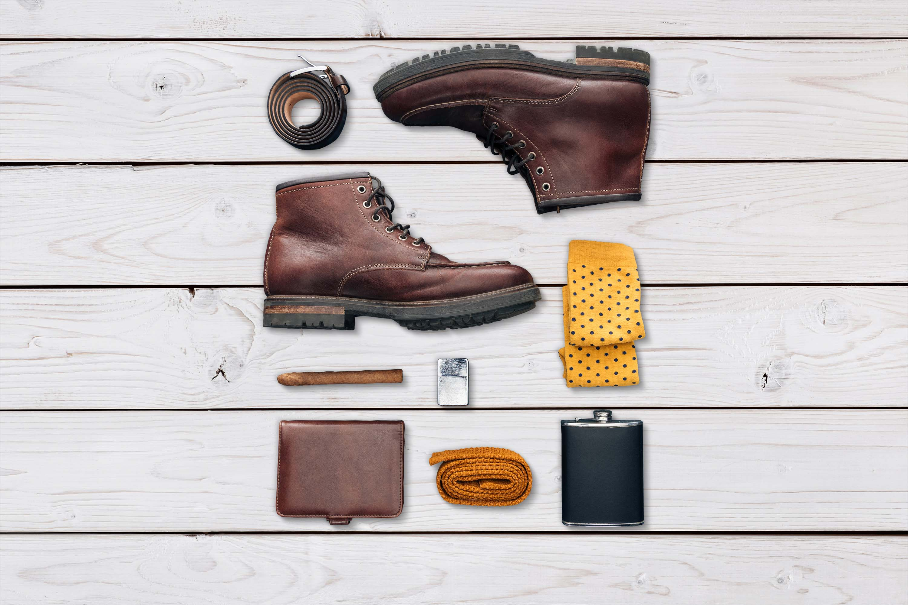 Herrenaccessoires auf einer weißen Holzoberfläche angeordnet: Stiefel, Gürtel, Socken und eine Flasche.