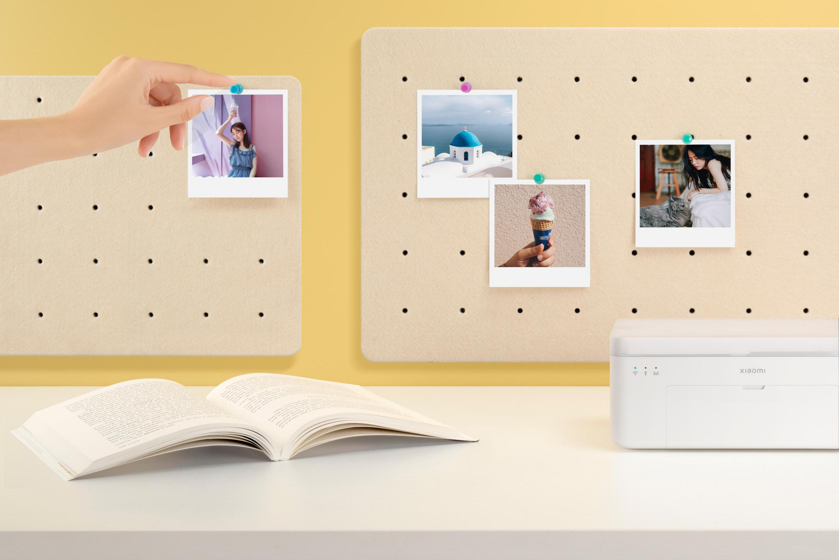 Ręka umieszczająca zdjęcie na tablicy korkowej z innymi zdjęciami i drukarką na żółto-białym biurku.