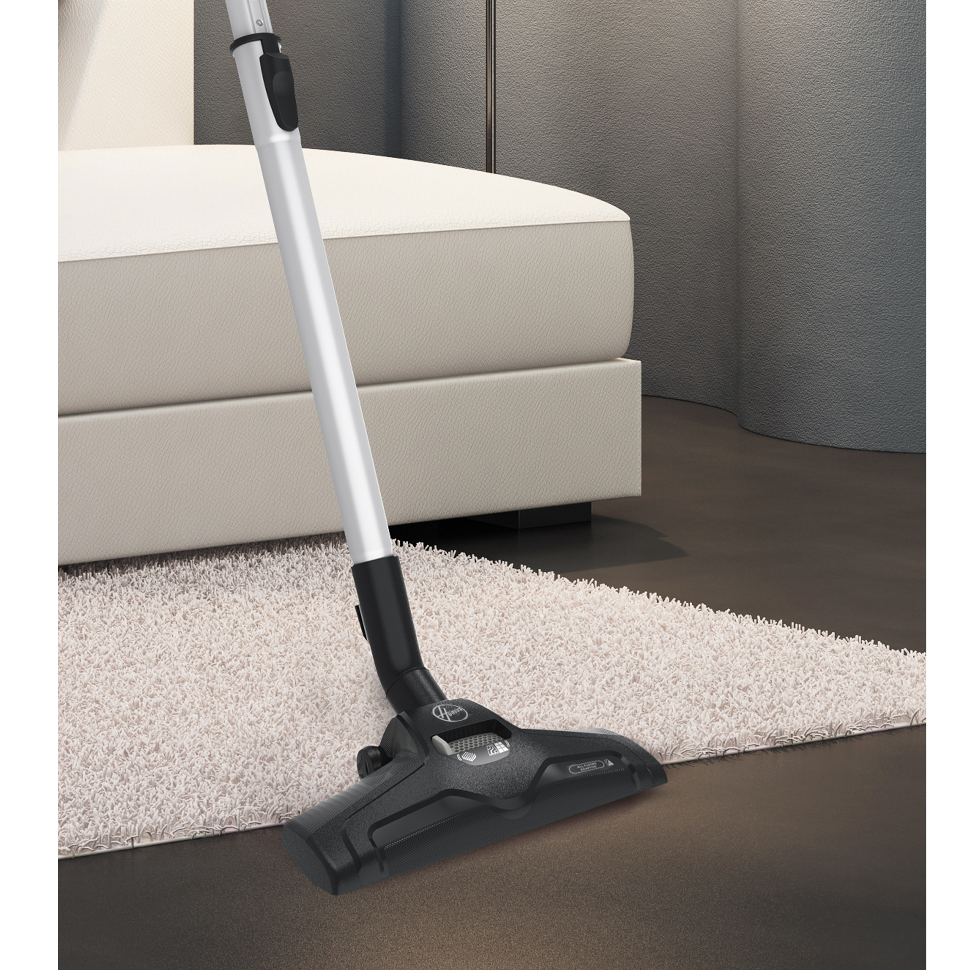 Odkurzacz ze srebrną rurą czyszczący jasnobeżowy dywan. W tle kanapa.