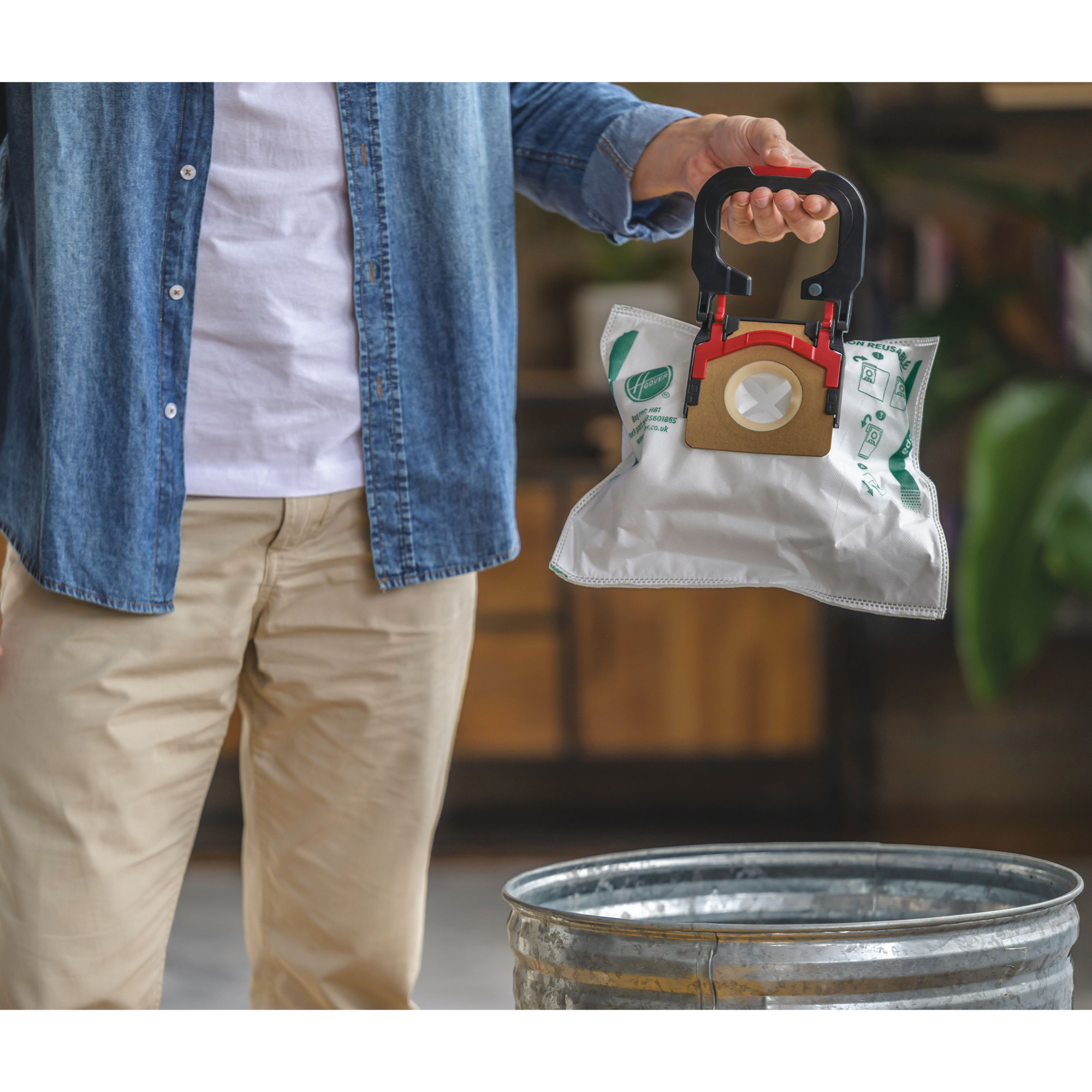 Osoba trzymająca worek do odkurzacza nad metalowym koszem na śmieci, z drewnianym wnętrzem w tle.