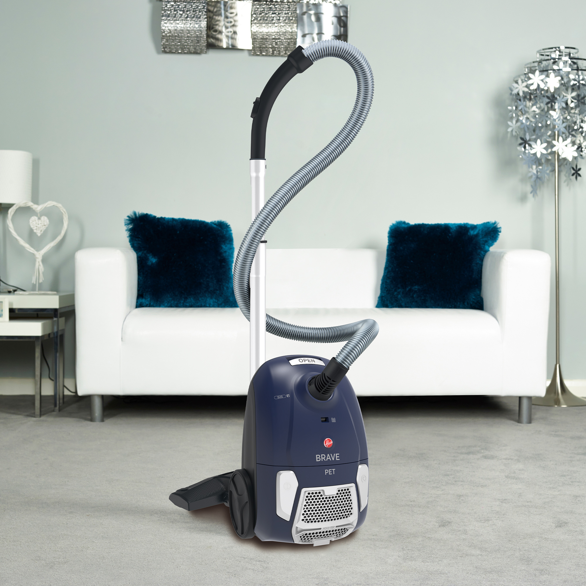 Niebieski odkurzacz na szarej wykładzinie. Biała sofa i niebieskie poduszki w tle.