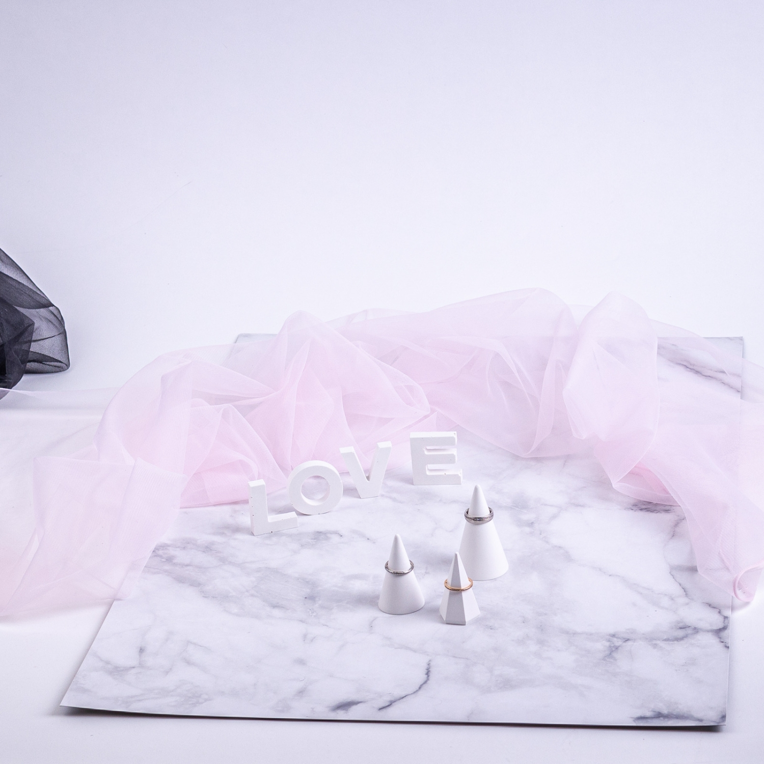 Weißer Hintergrund, rosa Stoff, weißer Buchstabe 'LOVE', weiße Ringhalter und silberne Ringe auf einer Marmoroberfläche.