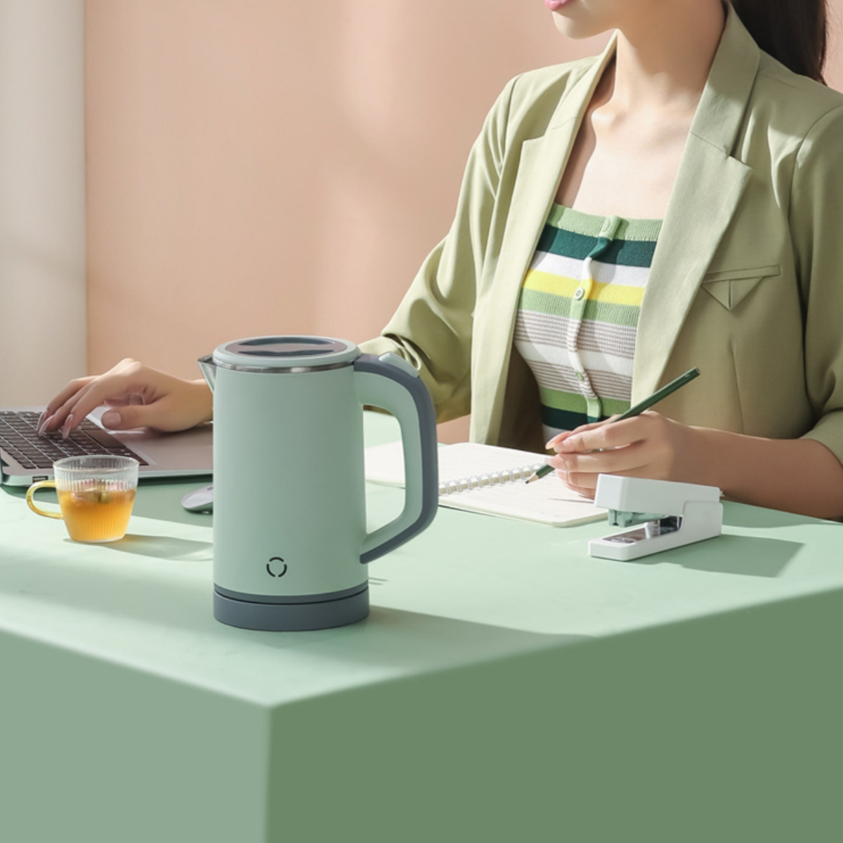 Eine Frau arbeitet an einem Schreibtisch mit Laptop, Wasserkocher und einer Tasse Tee.