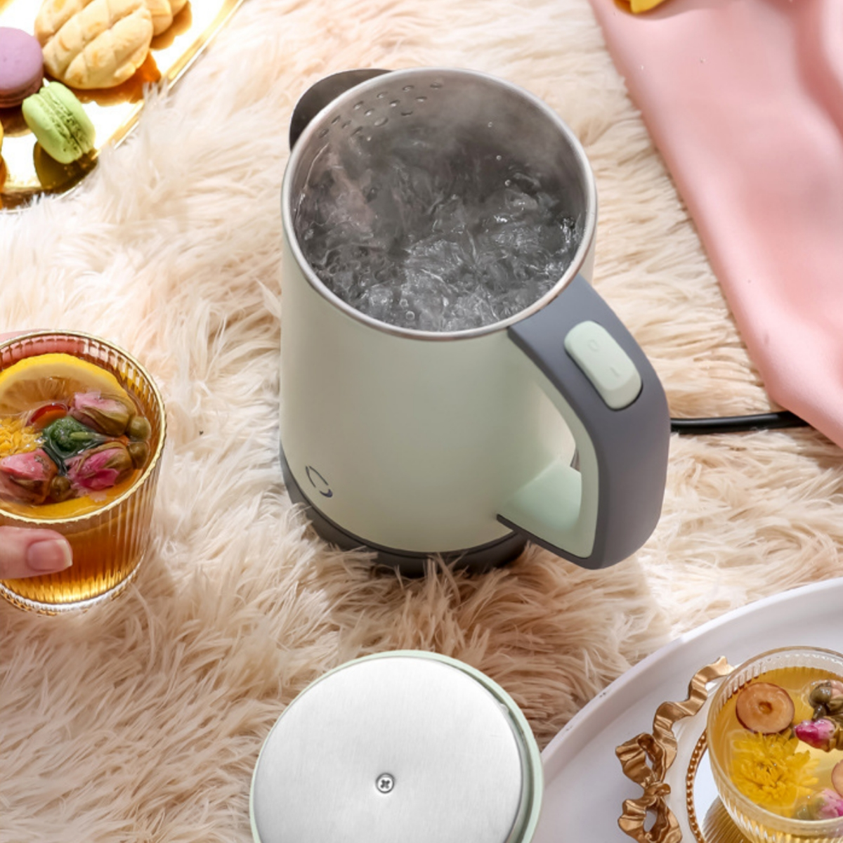 Kleiner grüner und grauer Wasserkocher mit kochendem Wasser, Tee und Makronen auf dem Tisch.
