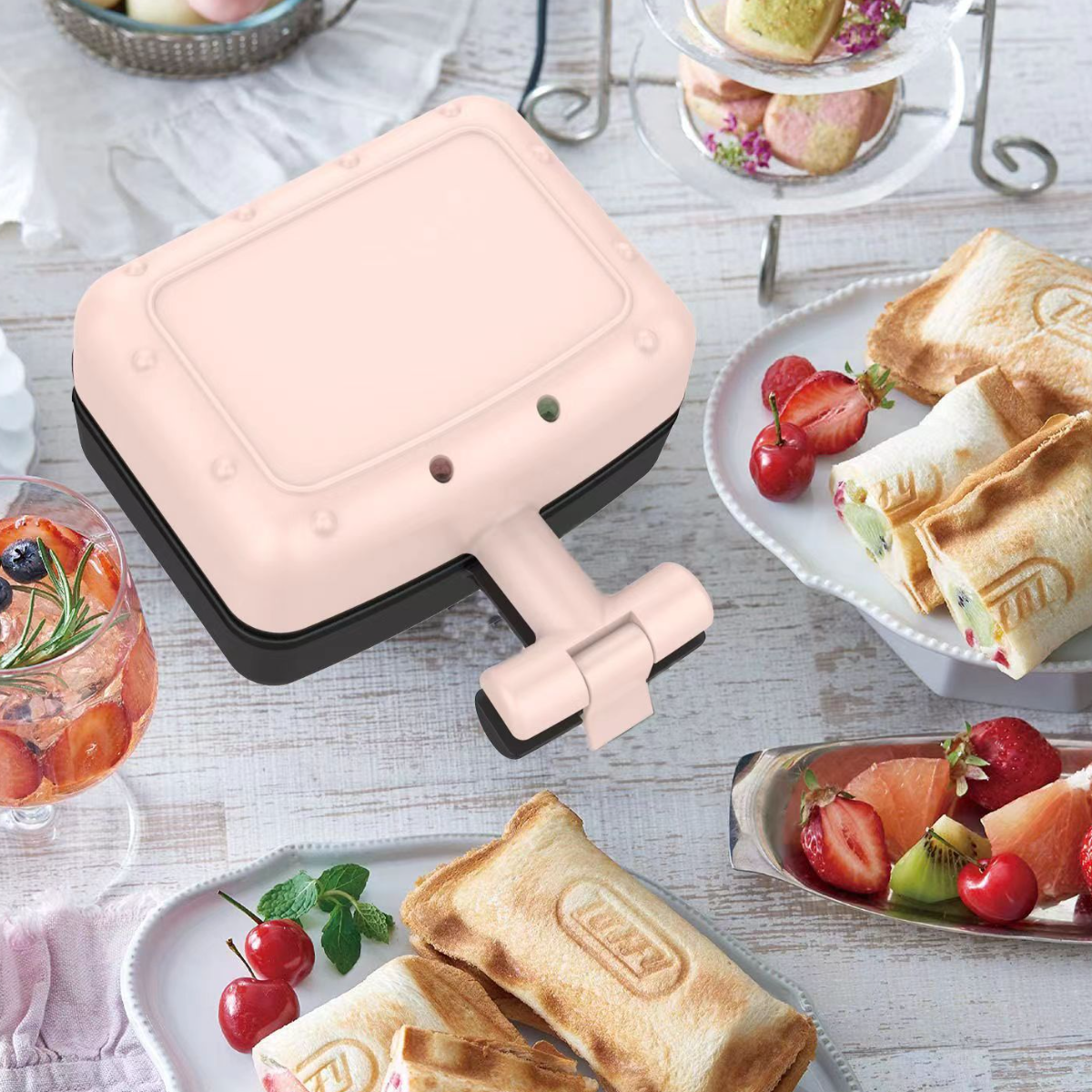 Rosa Waffeleisen mit Sandwiches und Obst auf einem weißen Holztisch.