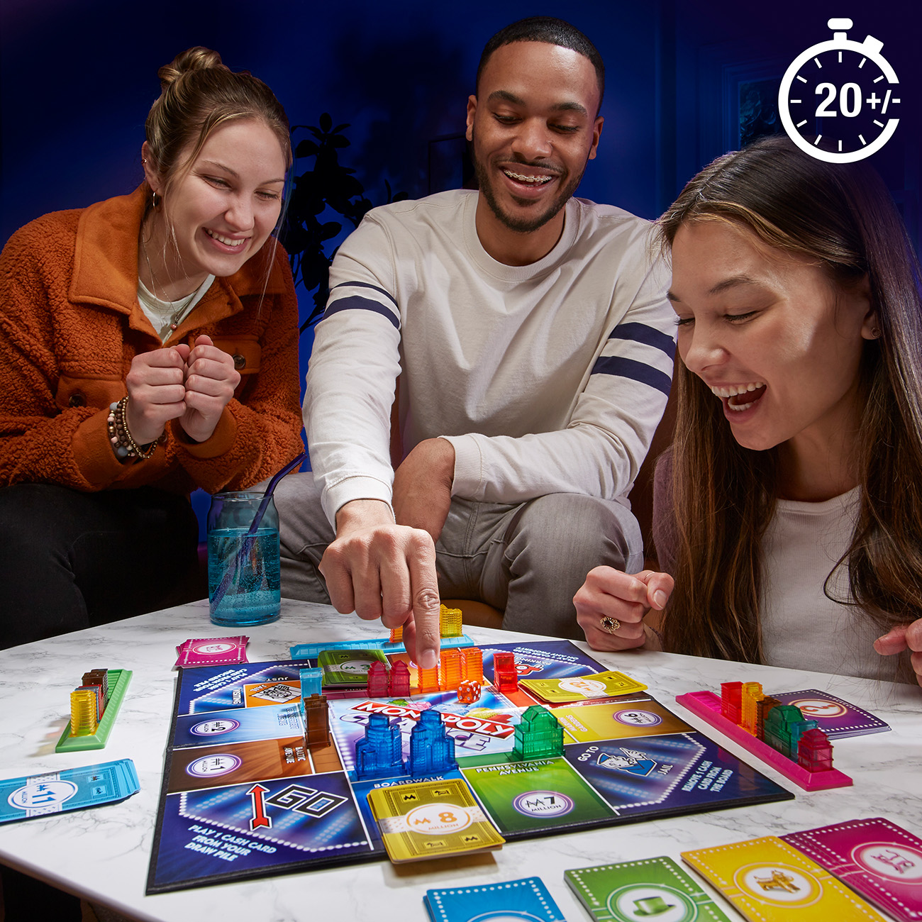 Drei Leute spielen Monopoly, mit Spielbrett, Grundstücken und Häusern. Der Timer zeigt 20+ Minuten.