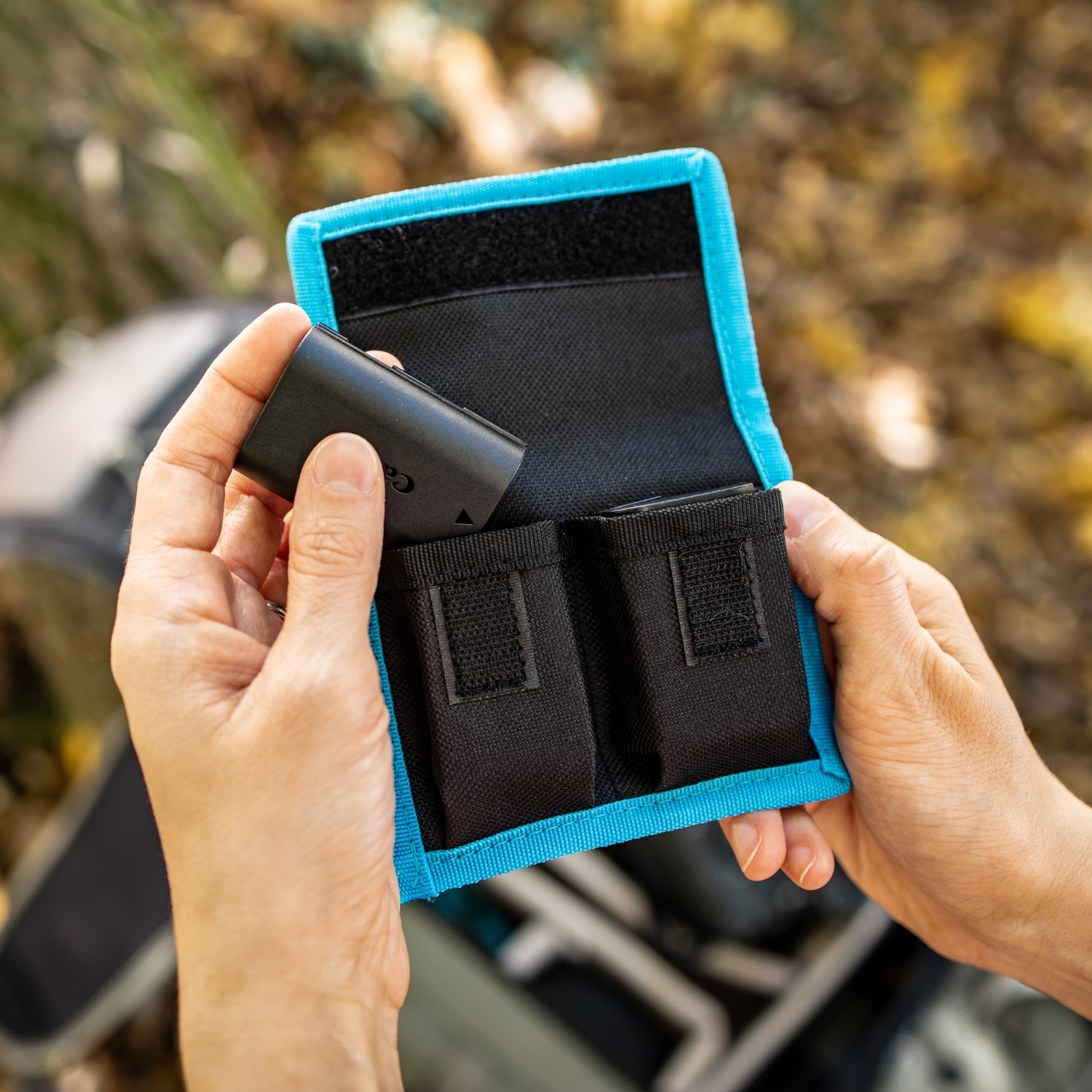 Hände halten eine schwarze Batterie und stecken sie in eine blau-schwarze Batterieaufbewahrungstasche.