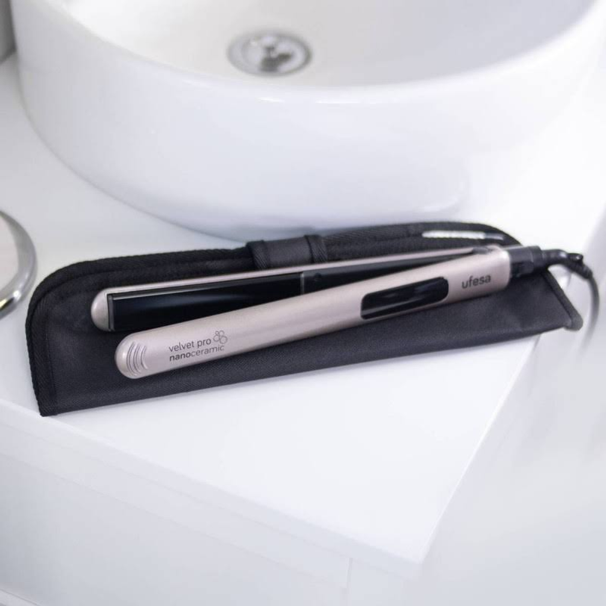 Plancha de pelo en un estuche negro sobre una superficie blanca junto a un lavabo blanco.