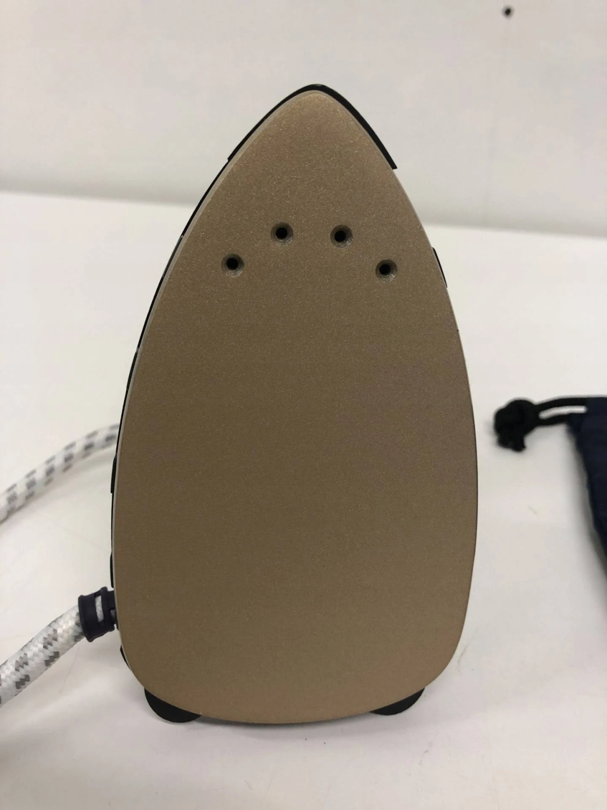Se ve la parte inferior de una plancha beige, con tres orificios de vapor y un borde negro, cable blanco.