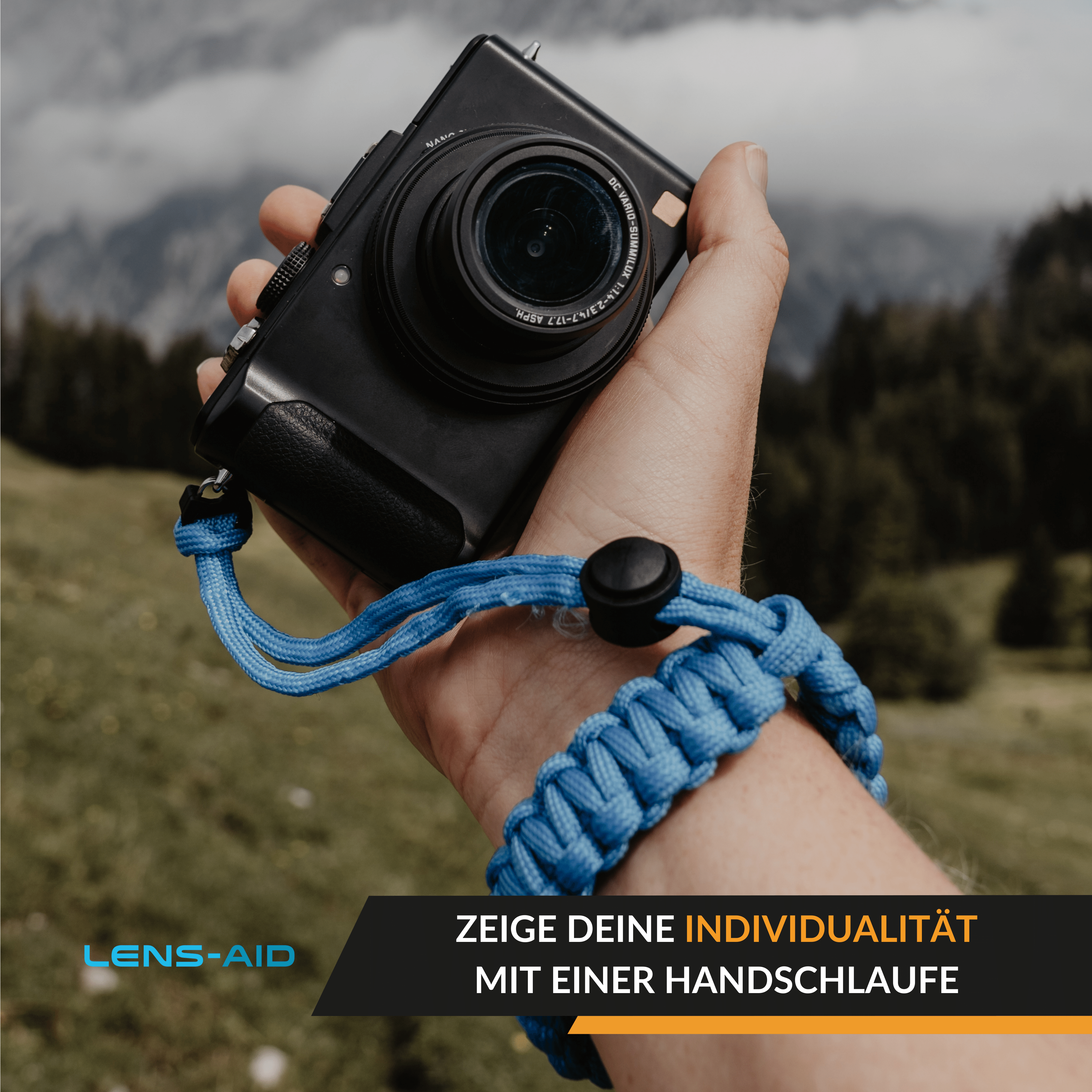 Mann hält eine schwarze Kamera mit blauem geflochtenen Handgelenkband. Berglandschaft im Hintergrund.