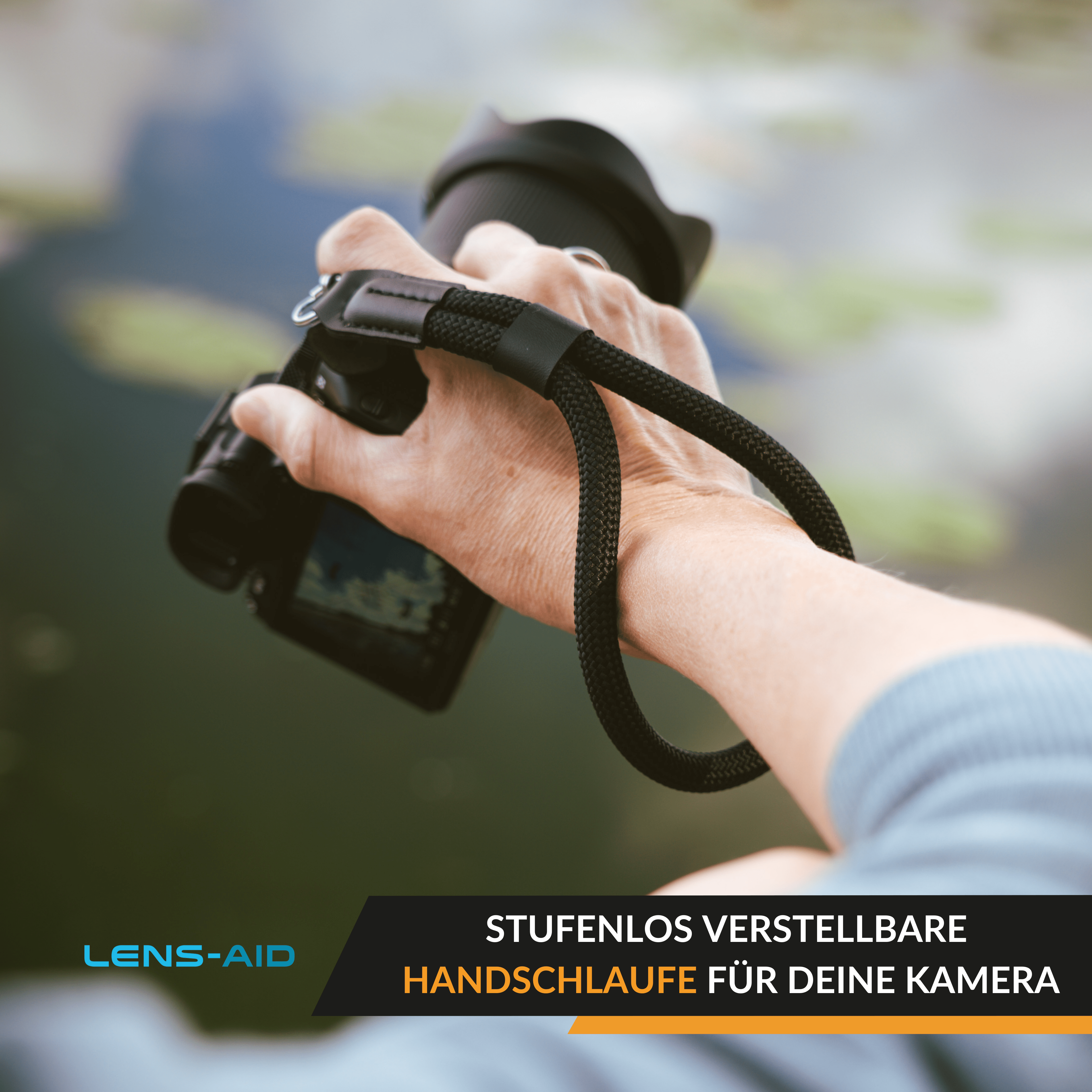 Hand hält eine schwarze Kamera mit schwarzem Gurt. Verschwommener Hintergrund mit Wasser und Pflanzen.