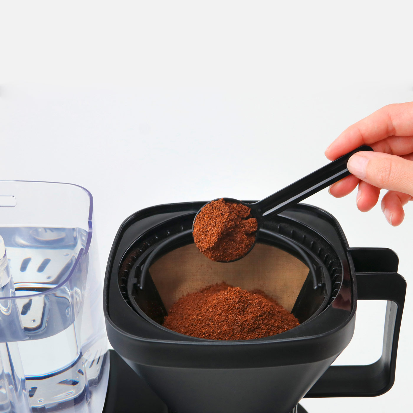 Eine Hand gießt Kaffeepulver in einen Kaffeefilter. Ein Wasserbehälter ist links sichtbar.
