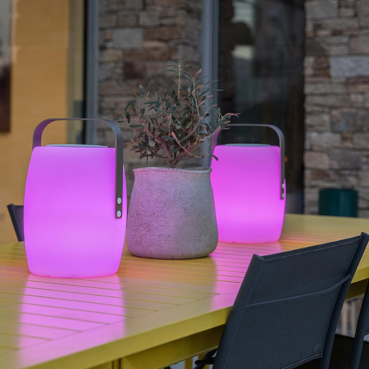 Zwei pink leuchtende Laternen, eine Topfpflanze und ein gelber Tisch auf einer Terrasse.