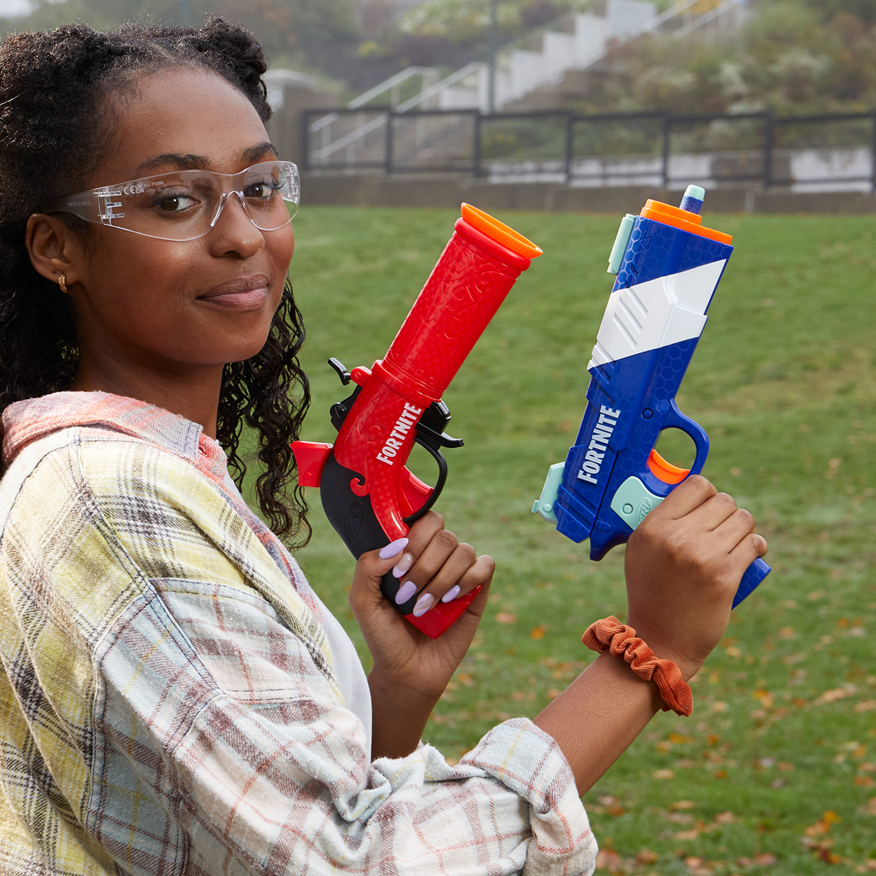 Frau mit Schutzbrille lächelt und hält zwei Fortnite-Spielzeugwaffen, eine rote und eine blaue, auf einem Feld.