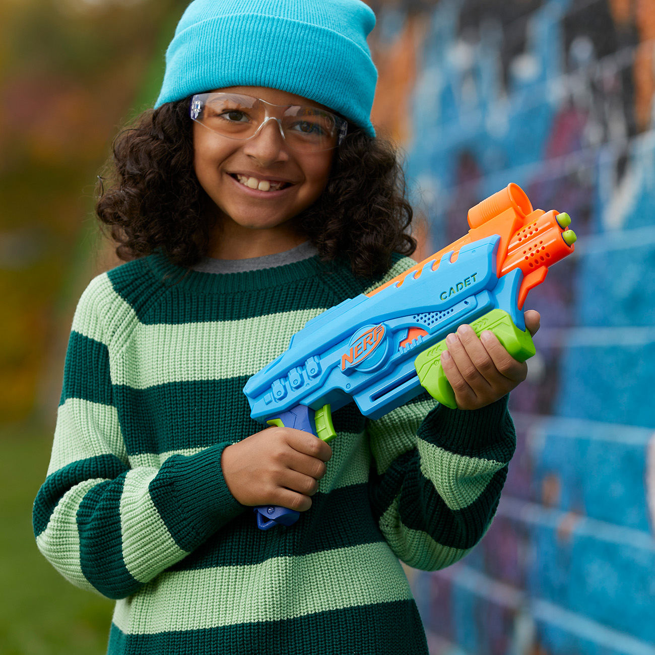 Bambino sorridente con cappello e occhiali blu che tiene in mano una pistola Nerf blu e arancione. Un muro di graffiti.