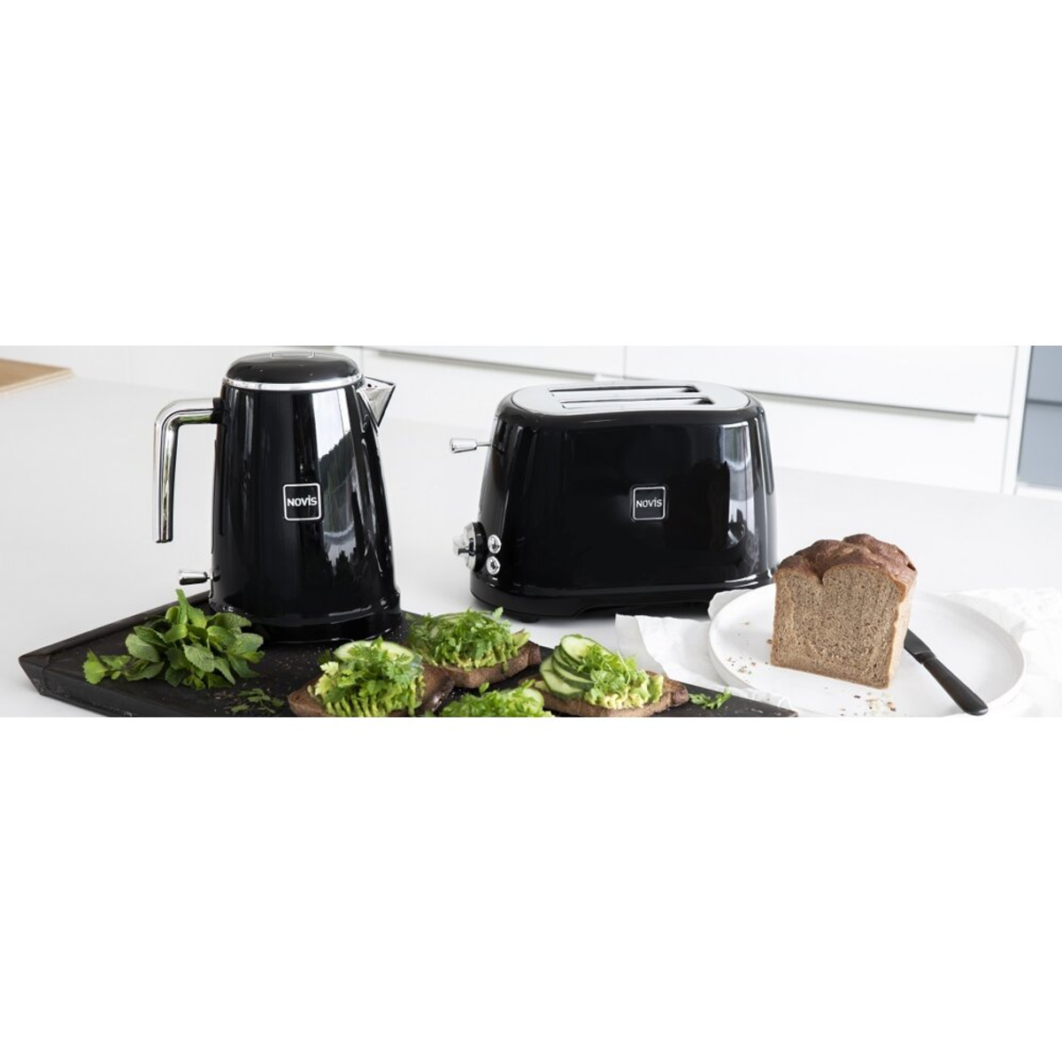 Schwarzer Wasserkocher und Toaster auf weißer Oberfläche mit Brot, Sandwiches und Kräutern im Vordergrund.