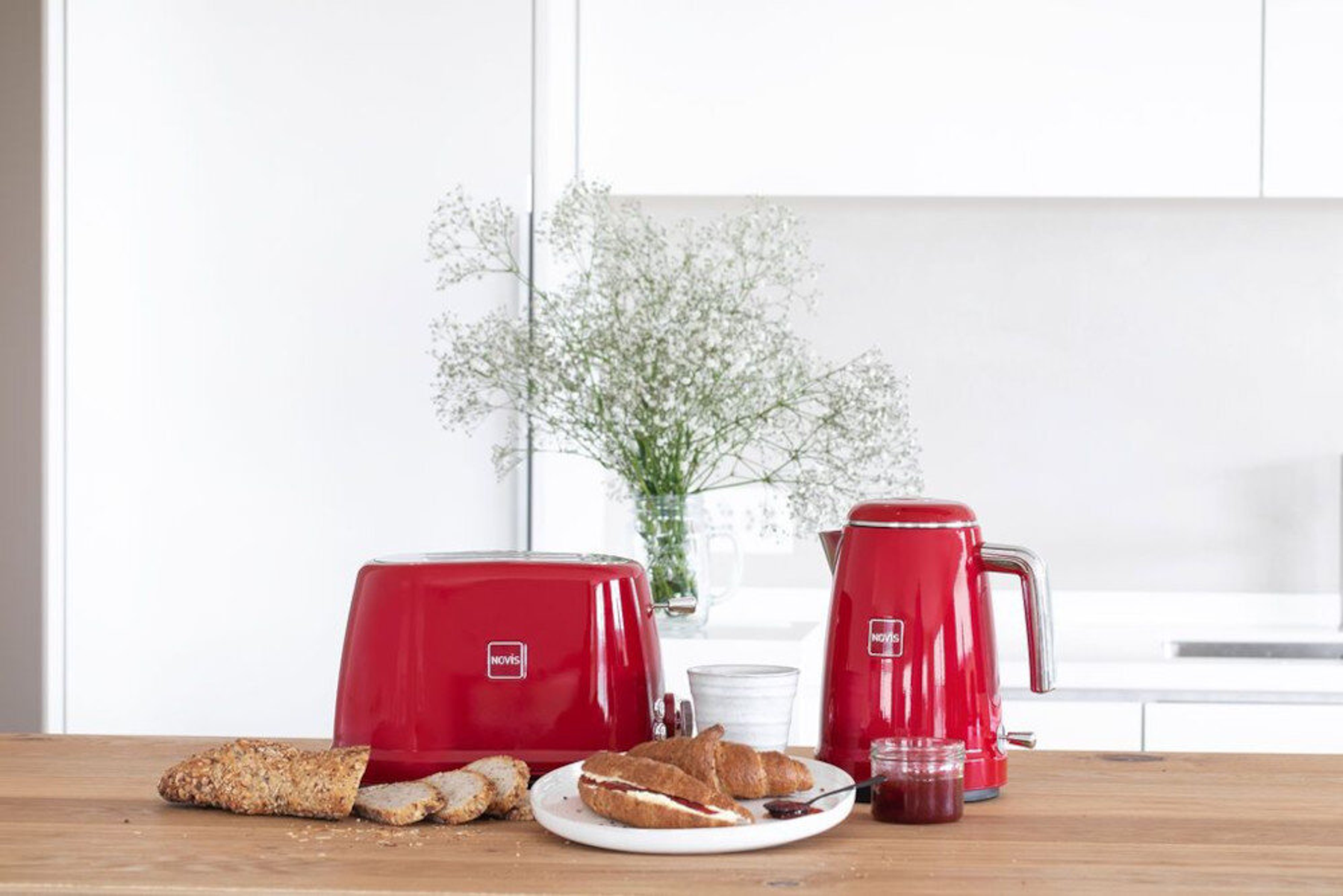Ein roter Toaster und Wasserkocher mit Croissants, Brot und Marmelade auf einem Holztisch mit Blumenvase.