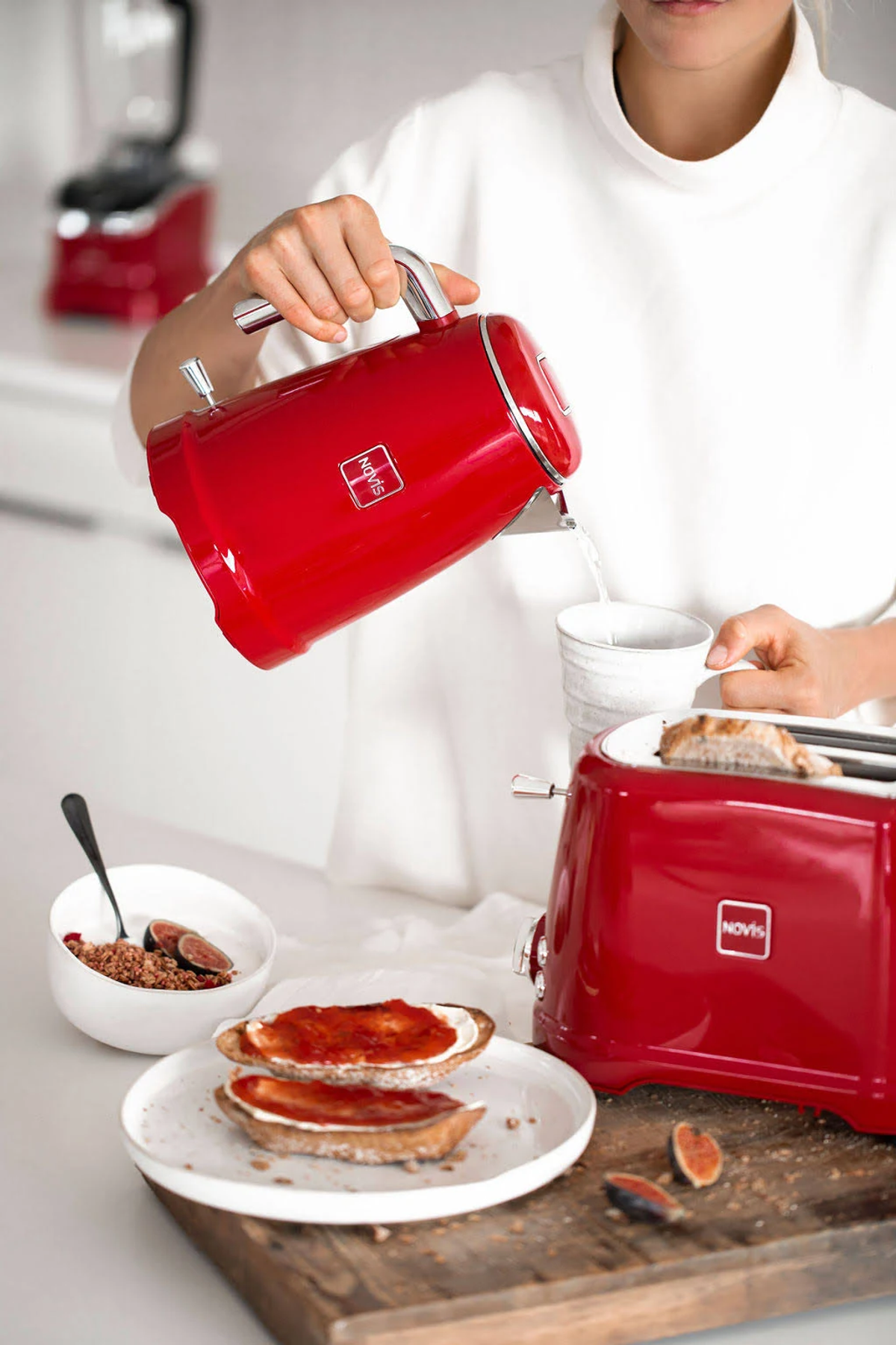 Donna che versa acqua da un bollitore elettrico rosso in una tazza bianca. Sono presenti altri oggetti.