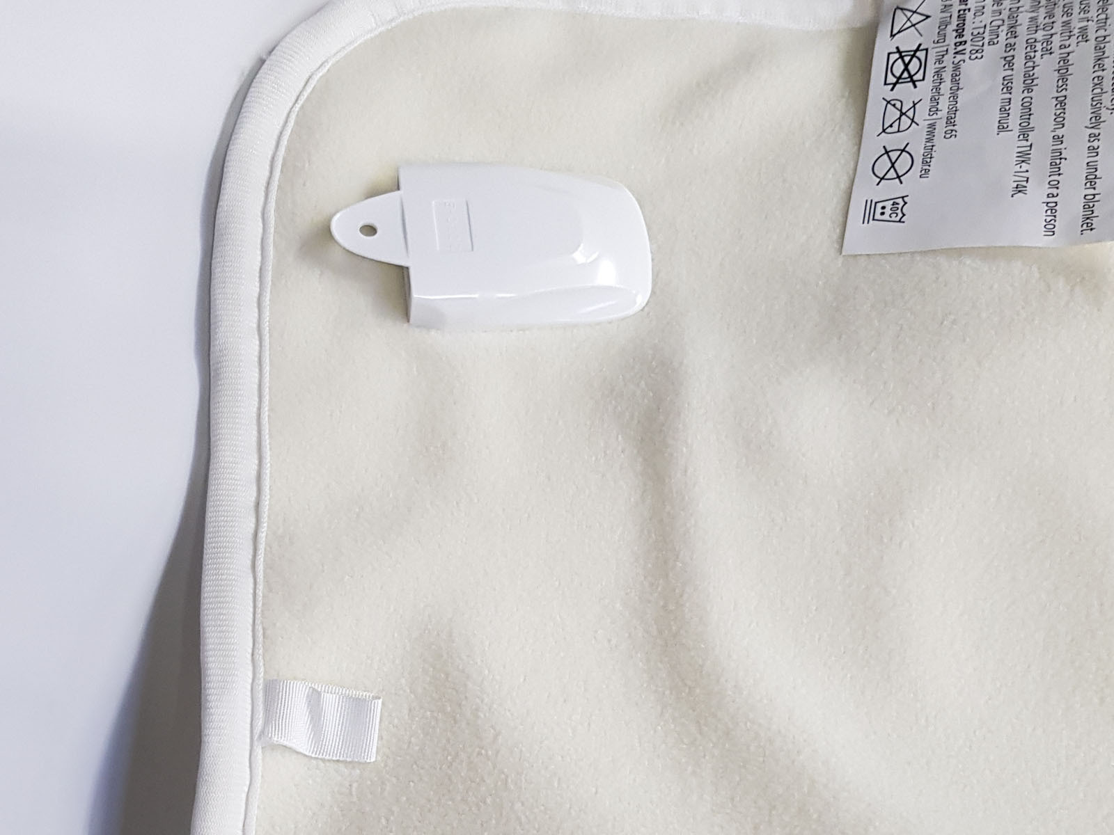 Biały koc elektryczny z białym plastikowym klipsem na białej powierzchni. Na kocu jest etykieta.