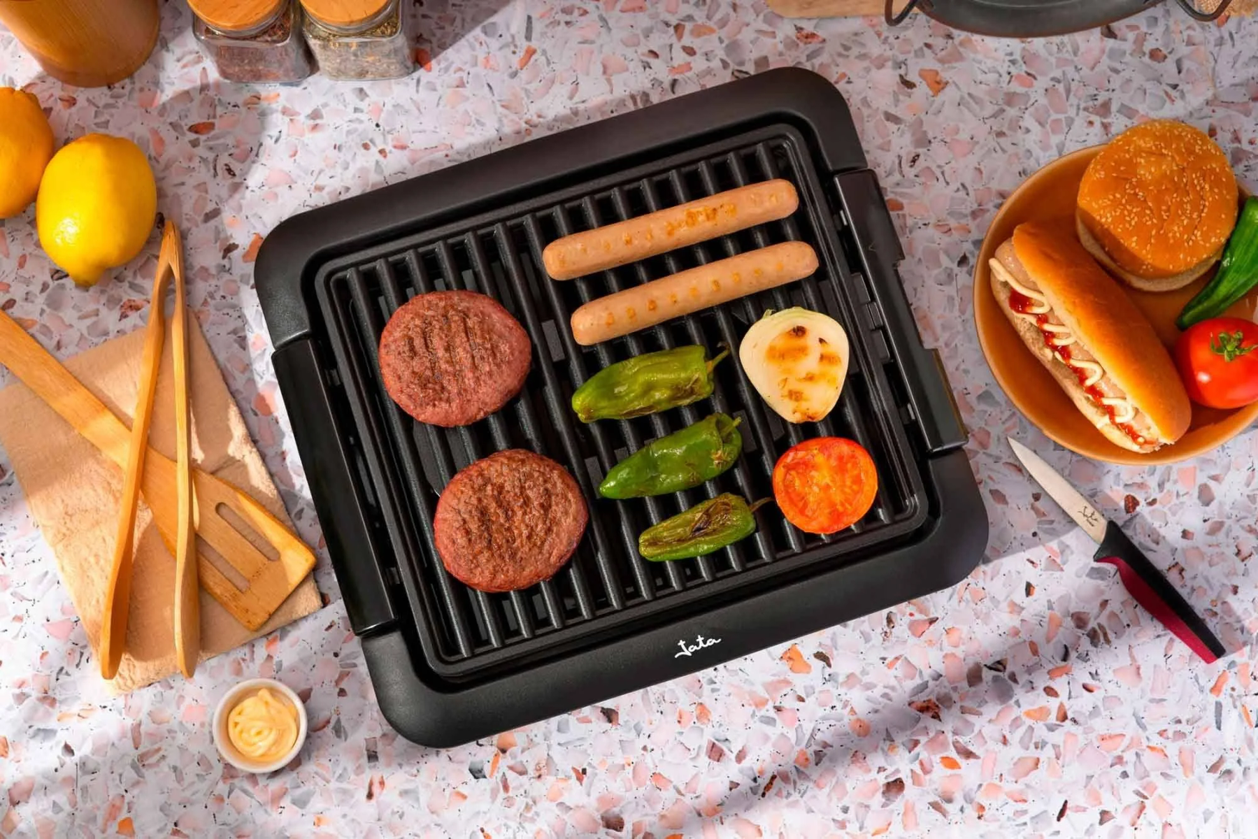 Una griglia nera con salsicce, hamburger, verdure grigliate e una salsa su un piano di lavoro.