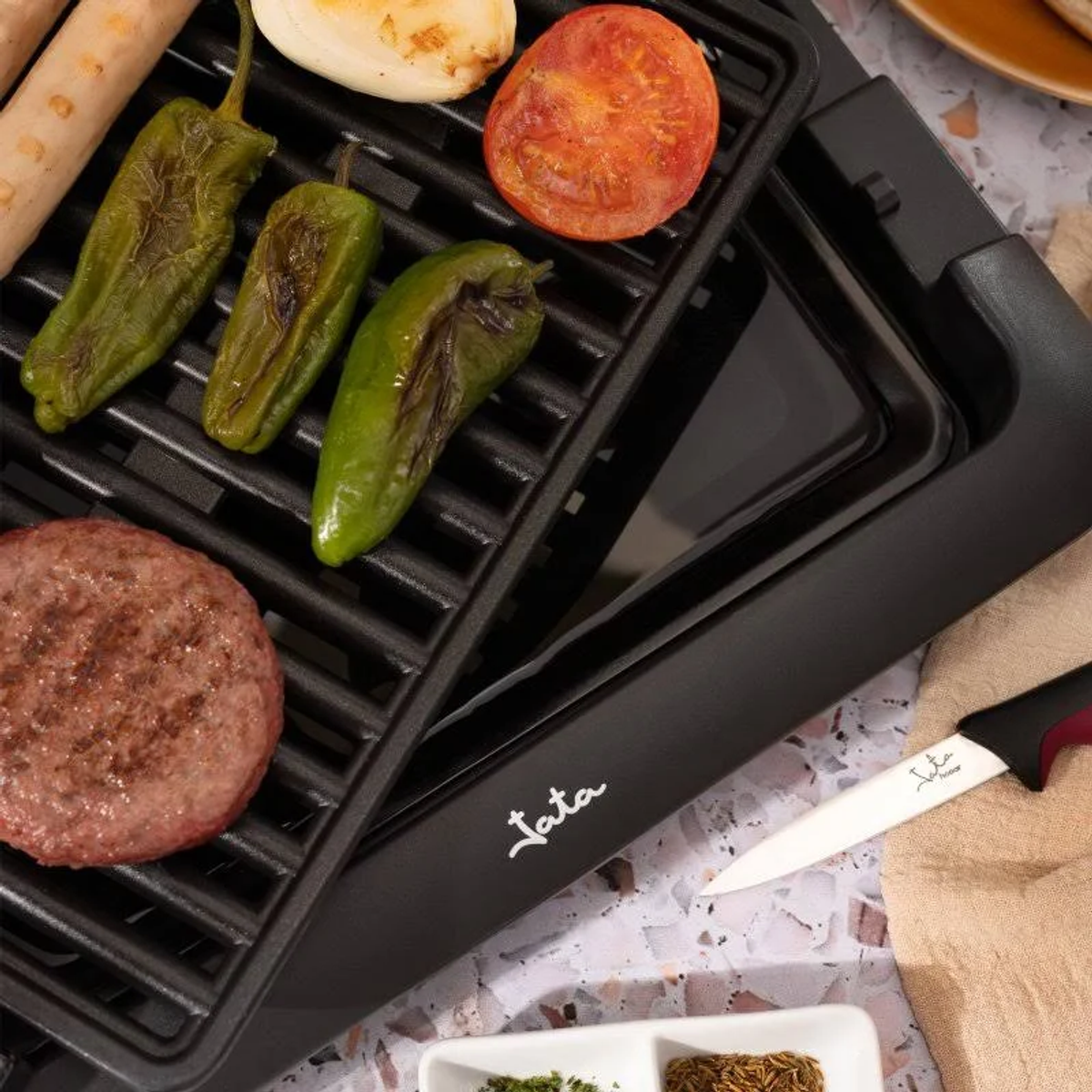 Una griglia con salsicce, peperoni, un pomodoro e un hamburger grigliati. Un coltello è di lato.