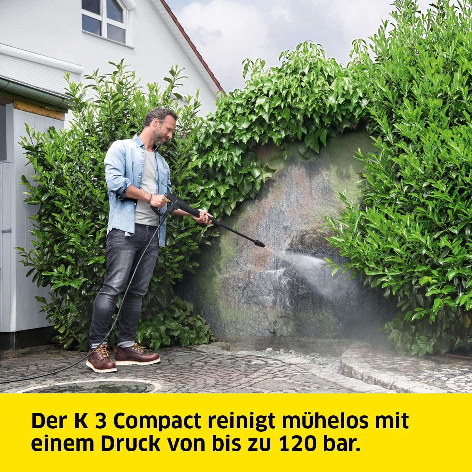 Mann reinigt mit einem Hochdruckreiniger eine Steinmauer mit dichtem Grün.
