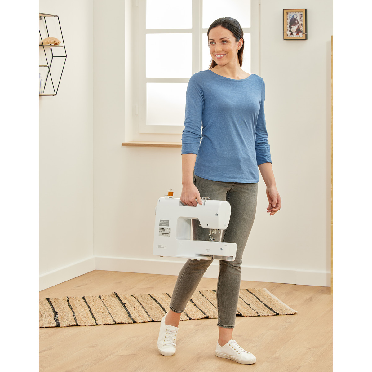 Kobieta trzymająca białą maszynę do szycia w pokoju z dywanem i oknem.