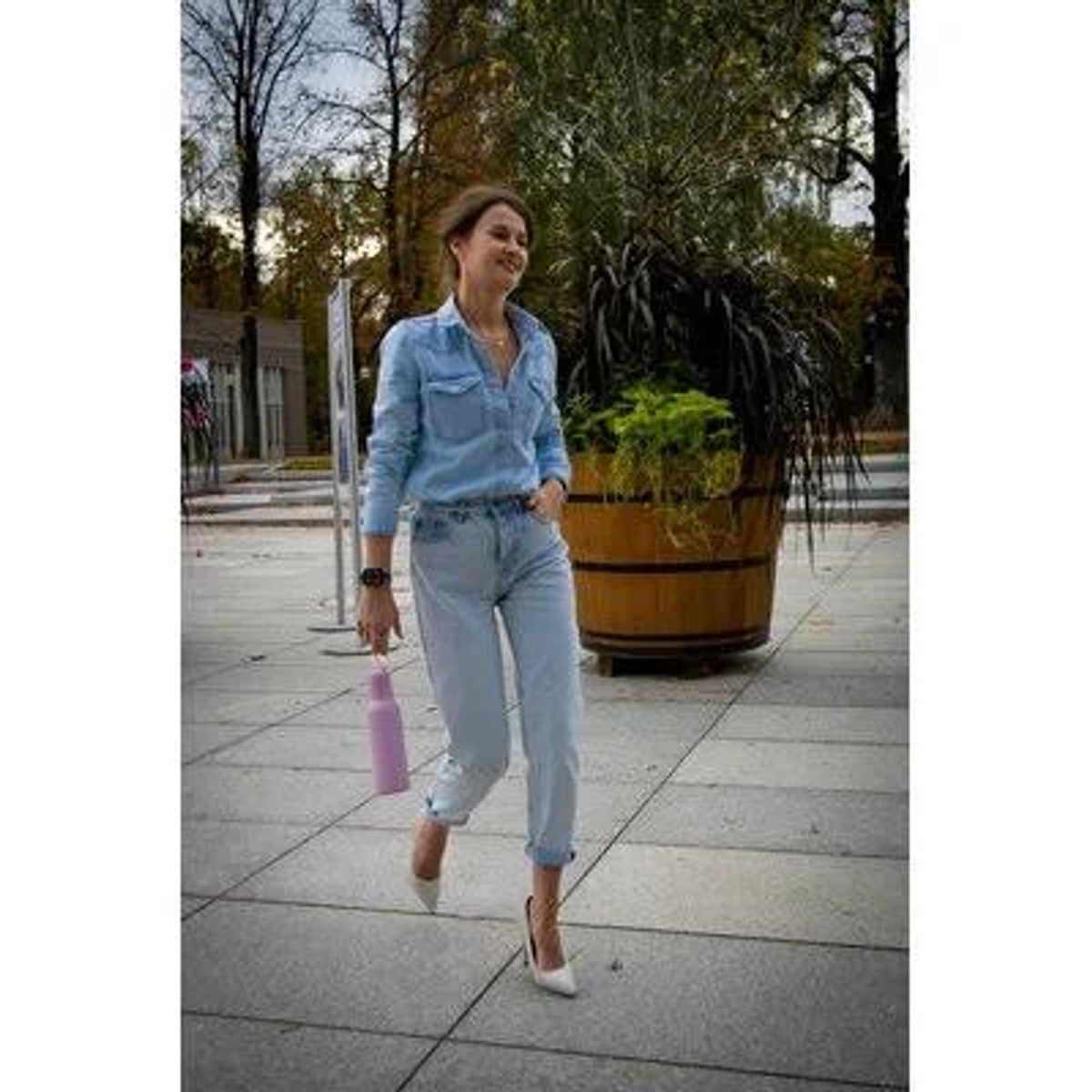 Kobieta idąca, trzymająca różową butelkę, w jasnych jeansach, szpilkach i koszuli dżinsowej.