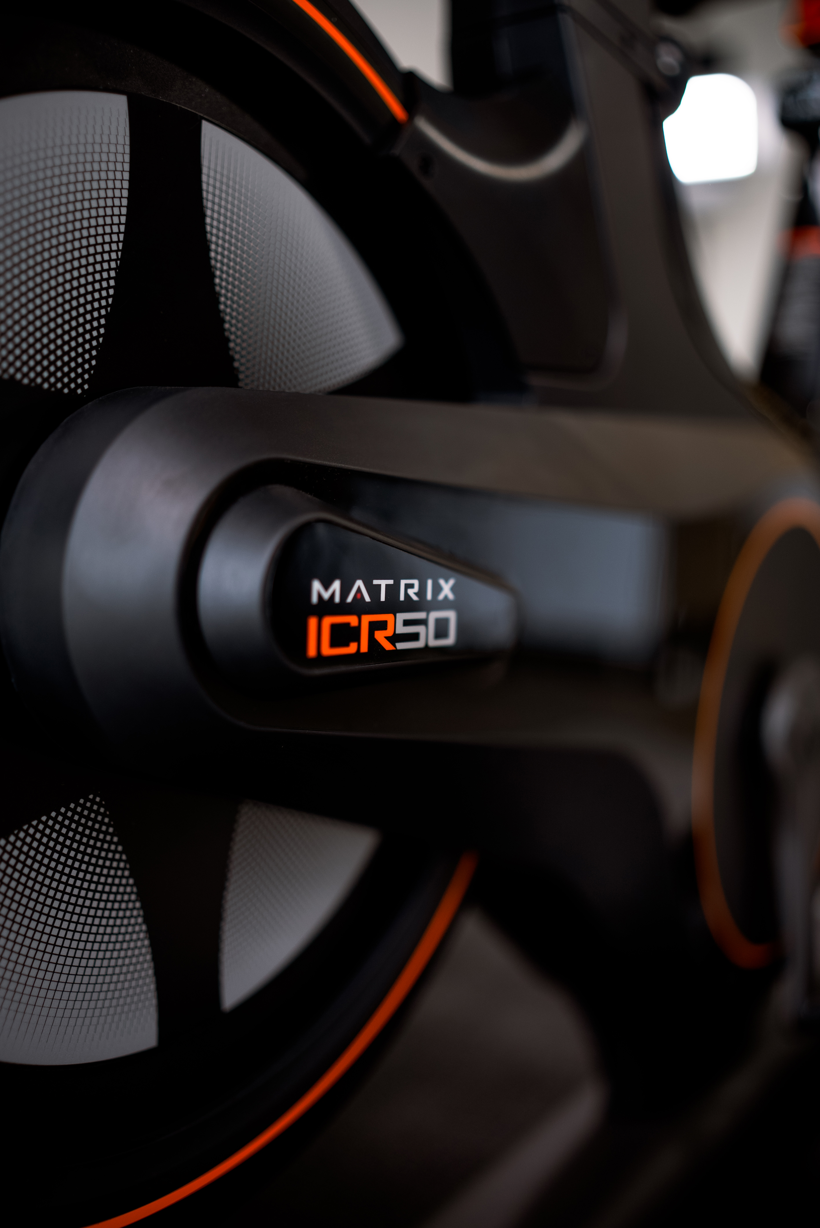 Détail d'un vélo d'exercice noir. Accents orange. Logo Matrix ICR50. Roues visibles. Fond flou.