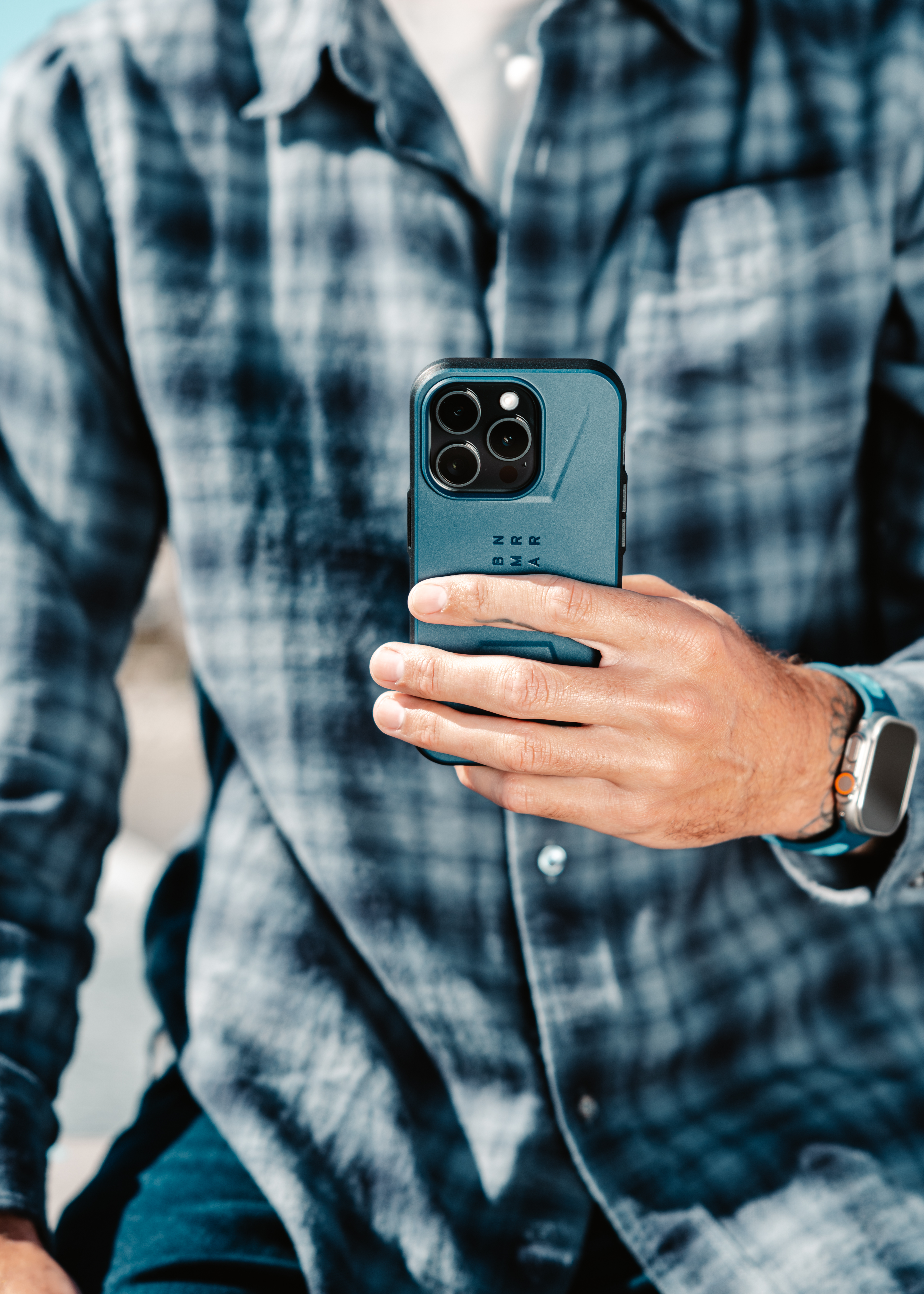 Ein Mann hält ein Telefon mit Schutzhülle, trägt ein kariertes Hemd und eine Uhr.
