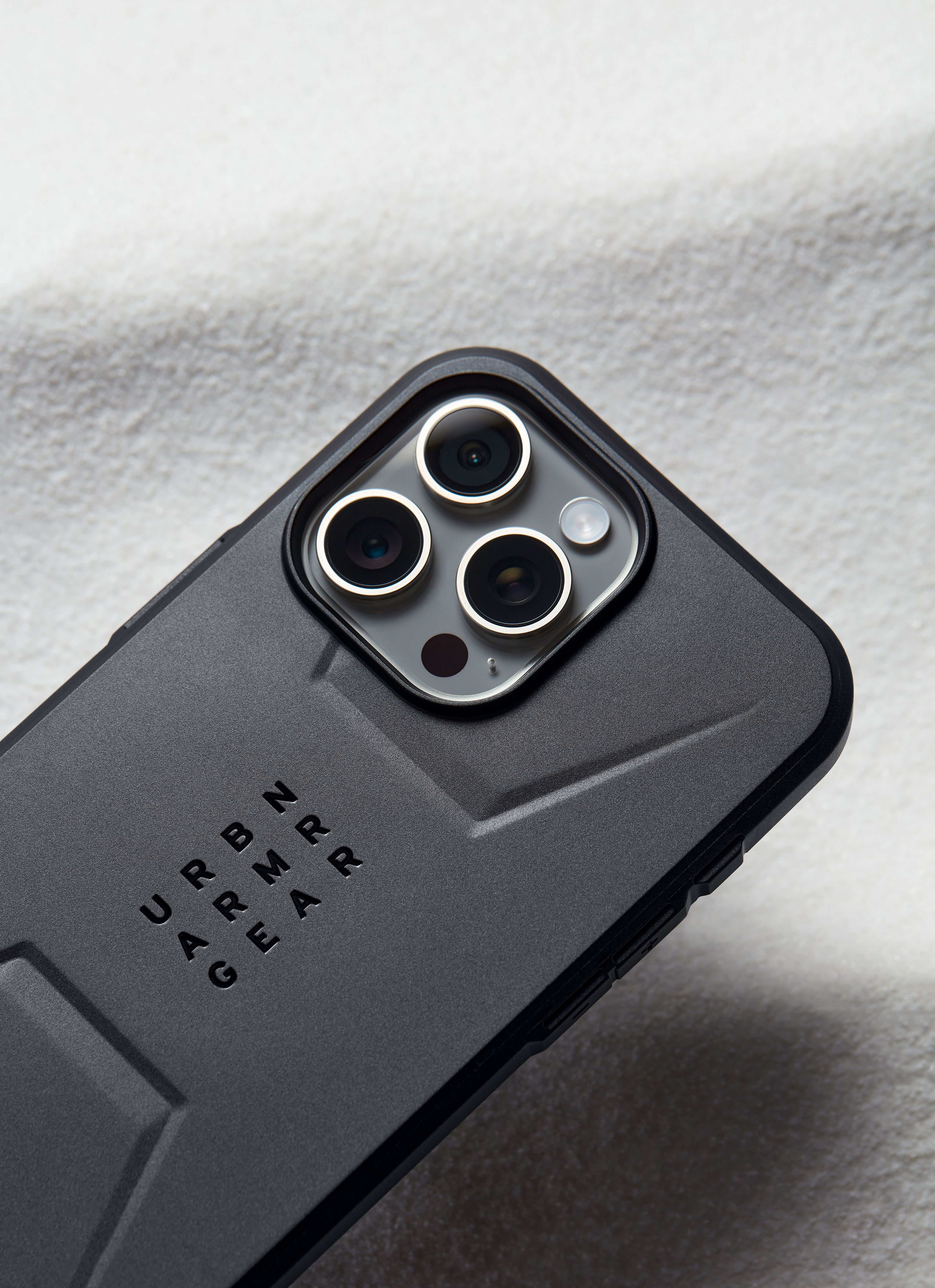 Funda de teléfono negra con texto 'URBAN ARMOR GEAR'. Tres lentes de cámara, fondo claro.