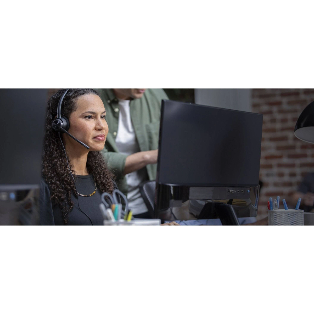 Vrouw met headset, kijkt naar een computerscherm, een andere persoon staat achter haar.