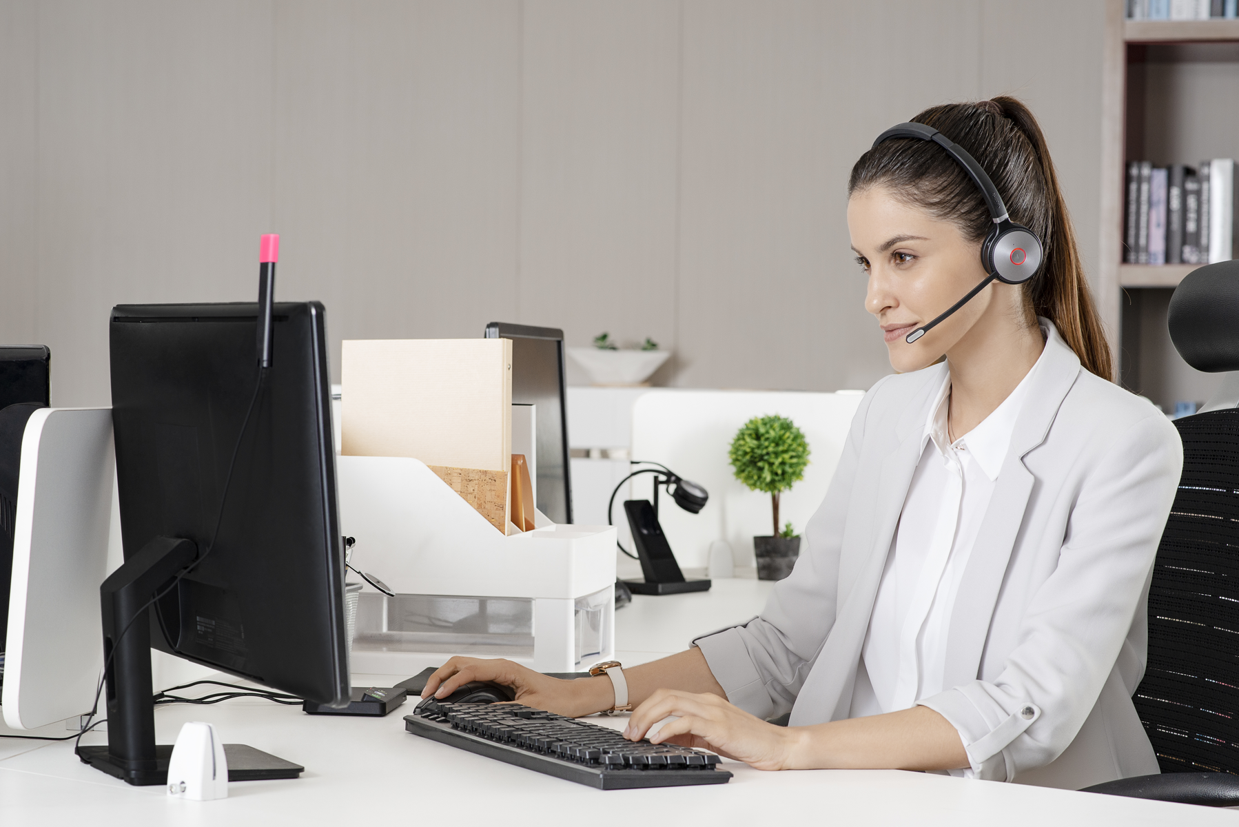 Frau benutzt Computer mit Headset. Schreibtisch hat Monitor, Tastatur und Bürobedarf.