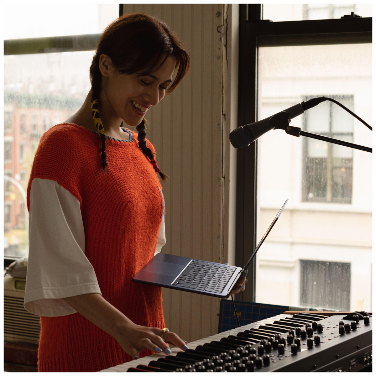 Frau spielt Keyboard. Sie hat Zöpfe und einen orangefarbenen Pullover. Ein Mikrofon im Hintergrund.