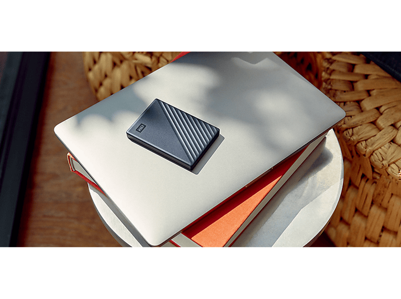 Laptop z dyskiem twardym na górze. Stos książek pod spodem, z wiklinowym koszem w tle.