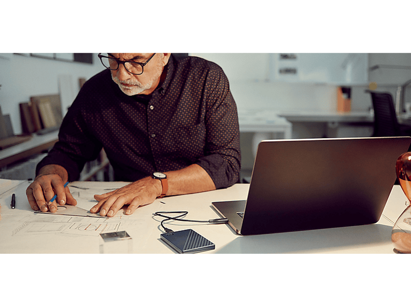 Mężczyzna pracuje przy biurku z laptopem, rysuje plany i ma dysk twardy.