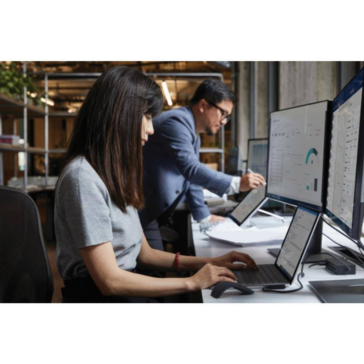 Dwie osoby pracujące w biurze, z wieloma ekranami komputerów wyświetlającymi wykresy i diagramy.