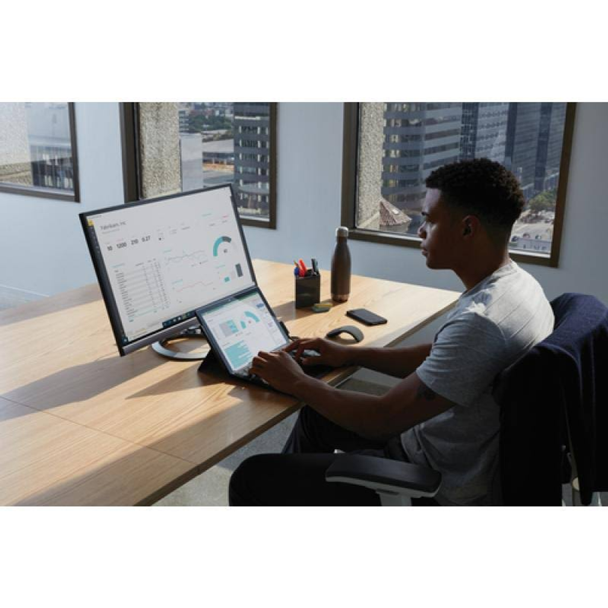 Mężczyzna korzystający z laptopa i monitora. Jest butelka wody i piórnik.