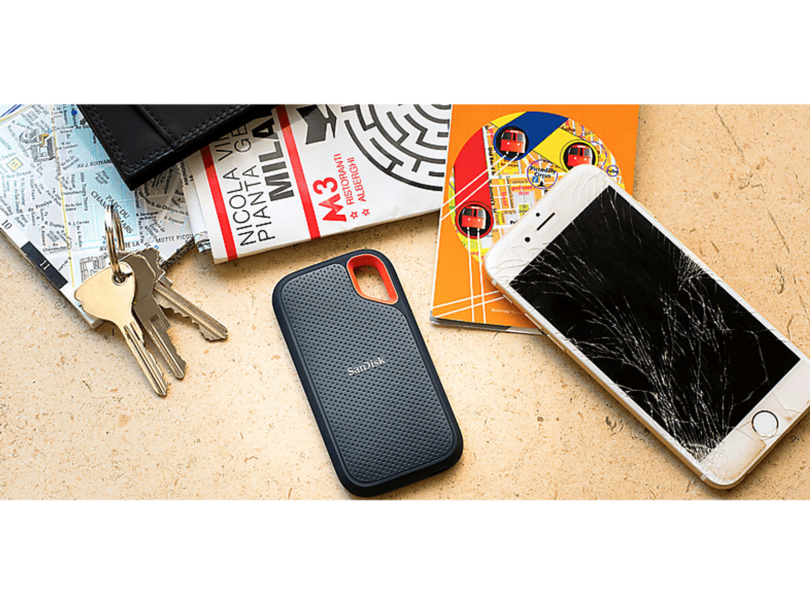 Telefon z pękniętym ekranem, klucze, portfel, zewnętrzny dysk twardy i mapa na beżowej powierzchni.