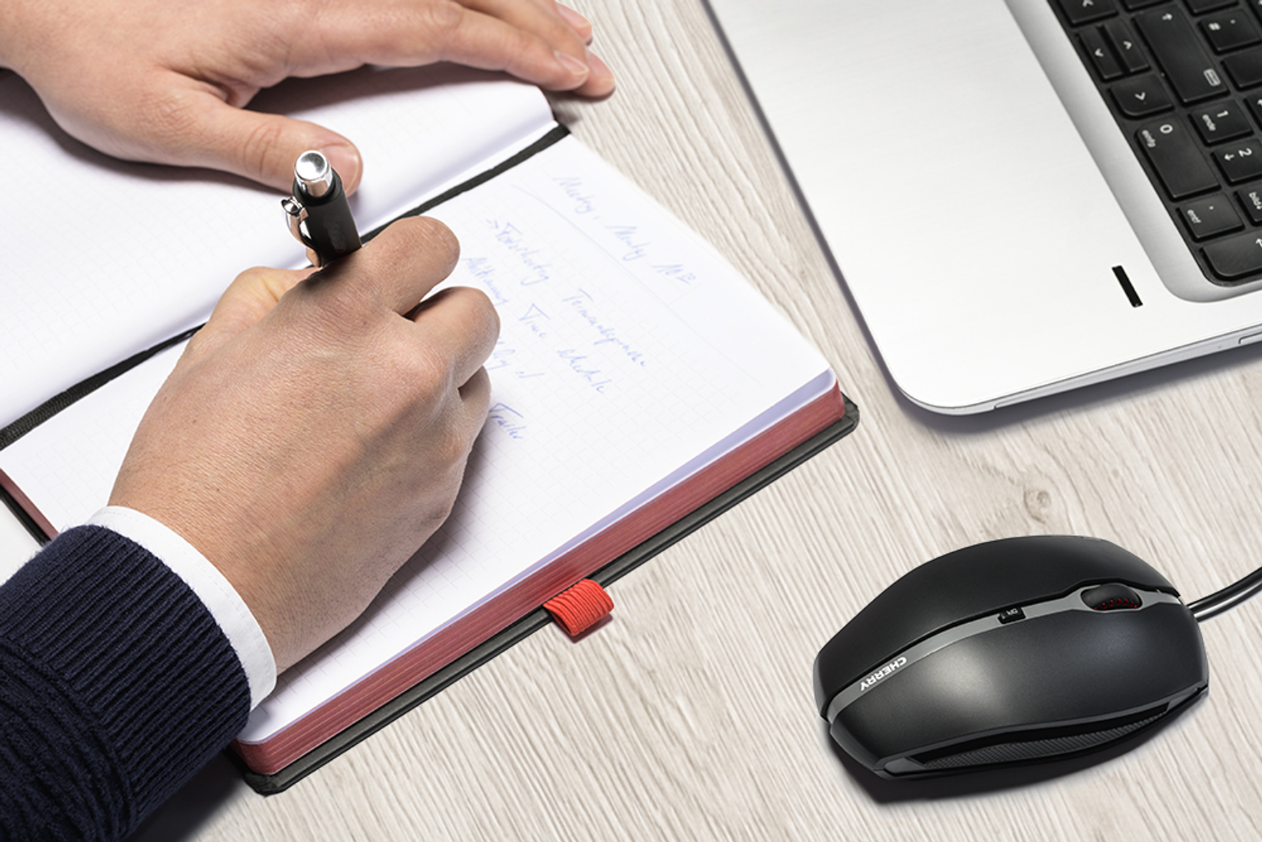 Manos escribiendo en un cuaderno junto a un portátil y un ratón Cherry negro en un escritorio de madera.