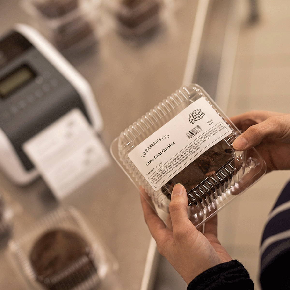 Dłonie trzymające przezroczysty plastikowy pojemnik z ciasteczkami. W tle drukarka etykiet.