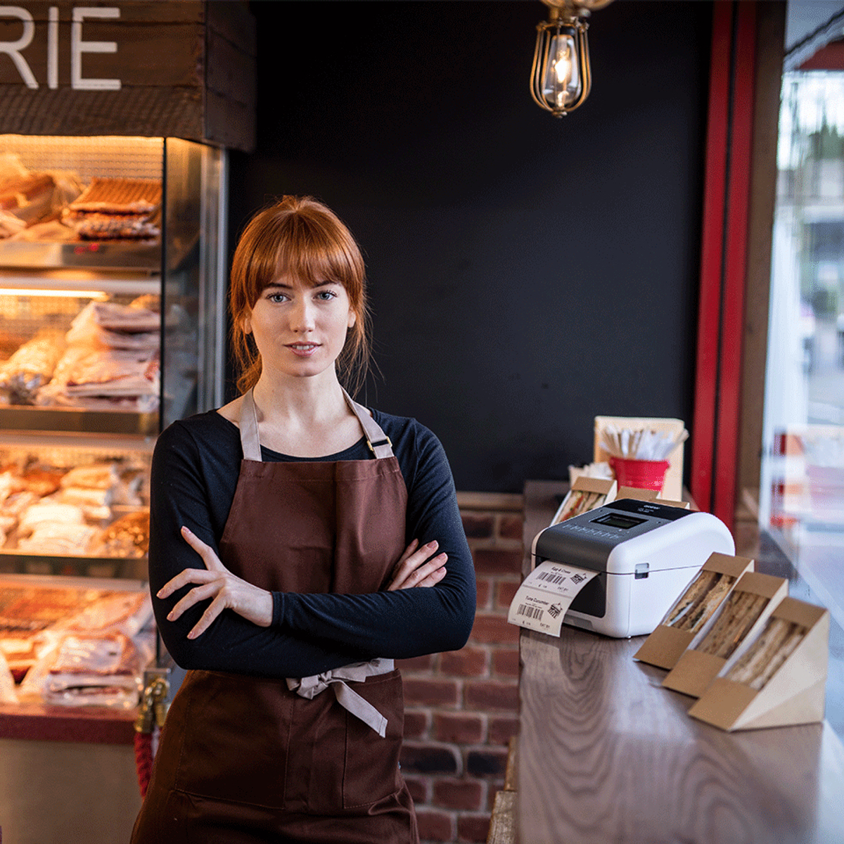 Kobieta w brązowym fartuchu, skrzyżowane ramiona, stoi w kawiarni. Za nią witryna.