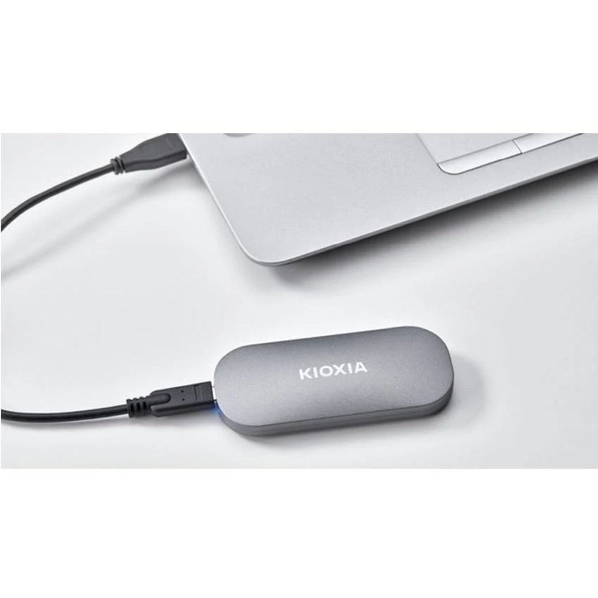 Srebrny zewnętrzny dysk twardy KIOXIA podłączony do laptopa za pomocą kabla USB, umieszczony na białej powierzchni.
